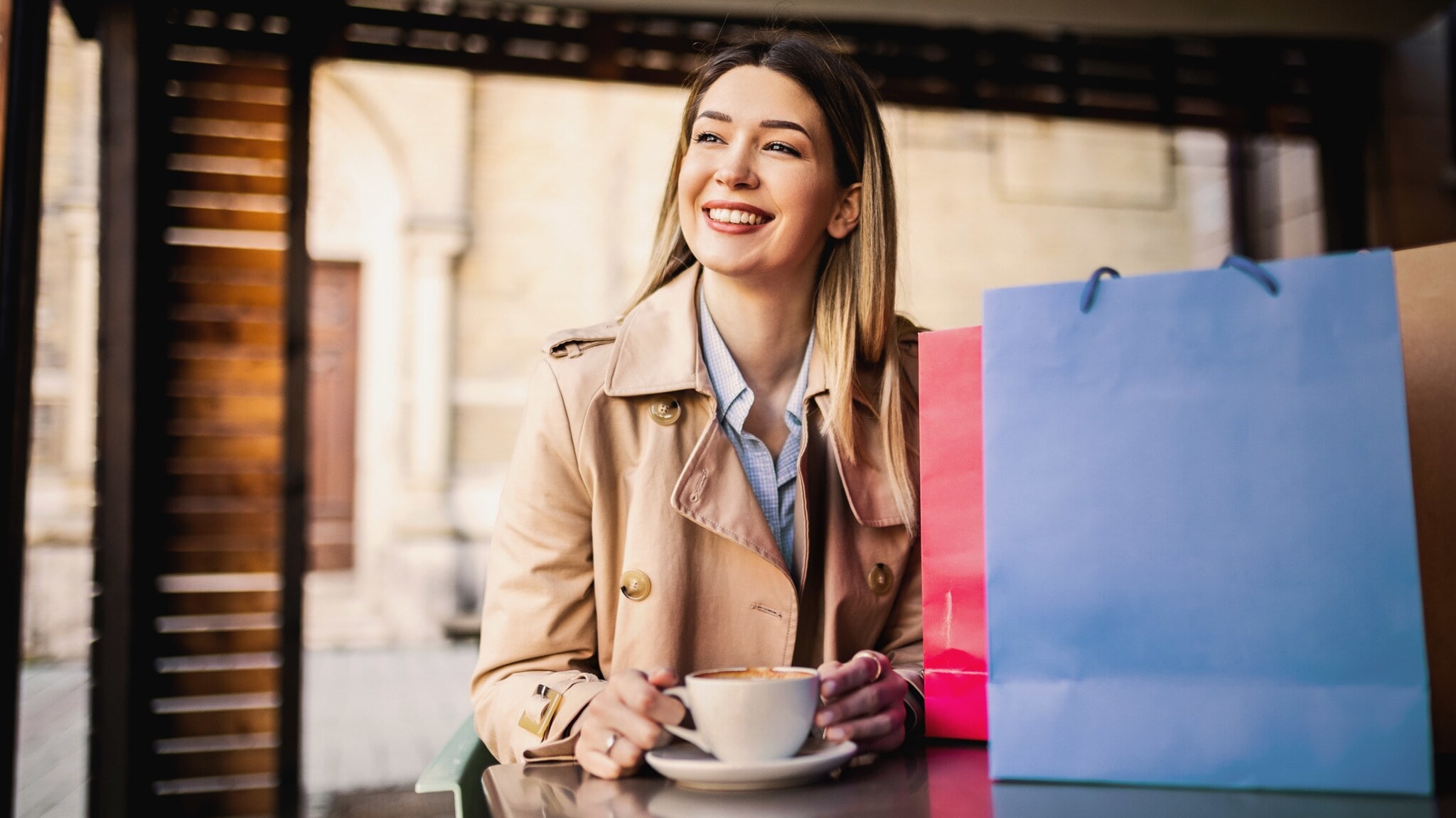 Eine Frau im Trenchcoat steht lächelnd mit einer Tasse Kaffee an einer Theke mit Einkaufstüten aus buntem Papier. Eine Frau im Trenchcoat steht lächelnd mit einer Tasse Kaffee an einer Theke mit Einkaufstüten aus buntem Papier.
