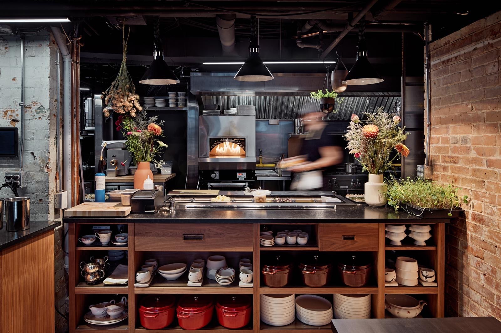 Blick in die Küche, wo im Ofen ein Feuer brennt und neben der unverputzten Backsteinwand eine Anrichte mit Blumensträußen und frischen Kräutern steht.