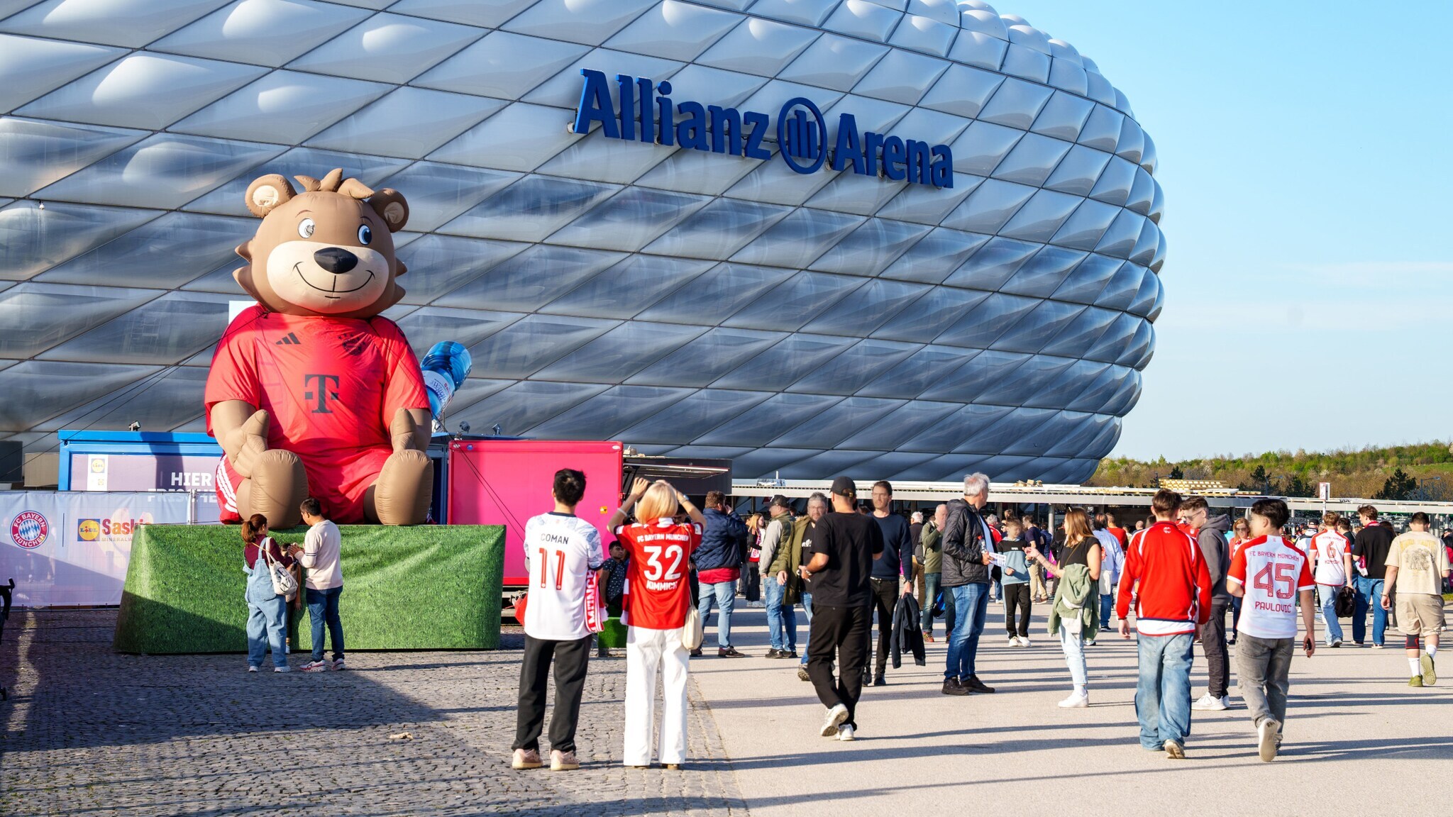 Personen mit Fußballtrikots am Eingang des Allianz Arena Stadion mit aufgeblasener Bärenfigur.