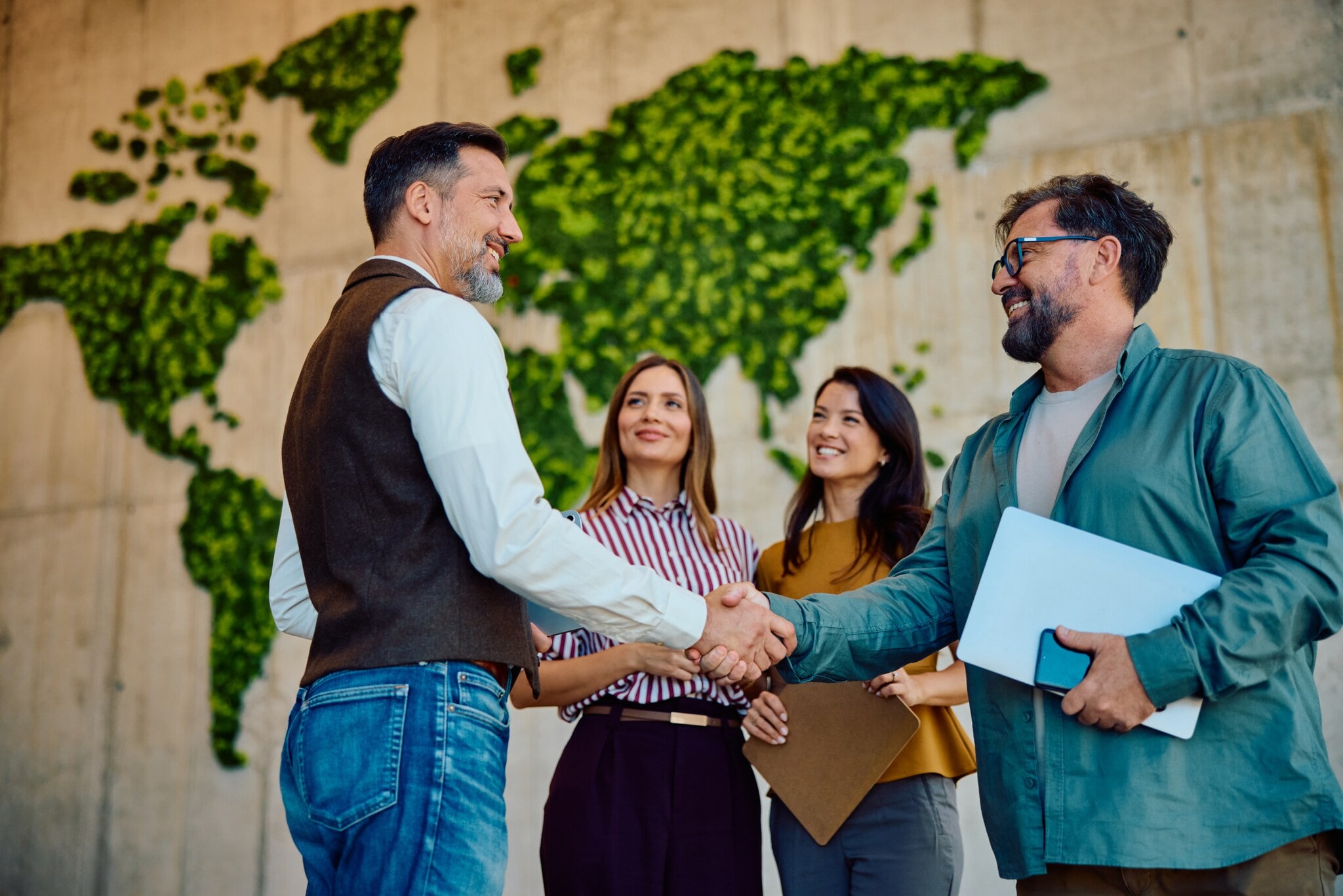 Zwei Männer mit Laptops im Arm geben sich lächelnd die Hand, daneben stehen zwei Frauen vor einer hohen Wand mit abstrakter, grüner Weltkarte. Zwei Männer mit Laptops im Arm geben sich lächelnd die Hand, daneben stehen zwei Frauen vor einer hohen Wand mit abstrakter, grüner Weltkarte.
