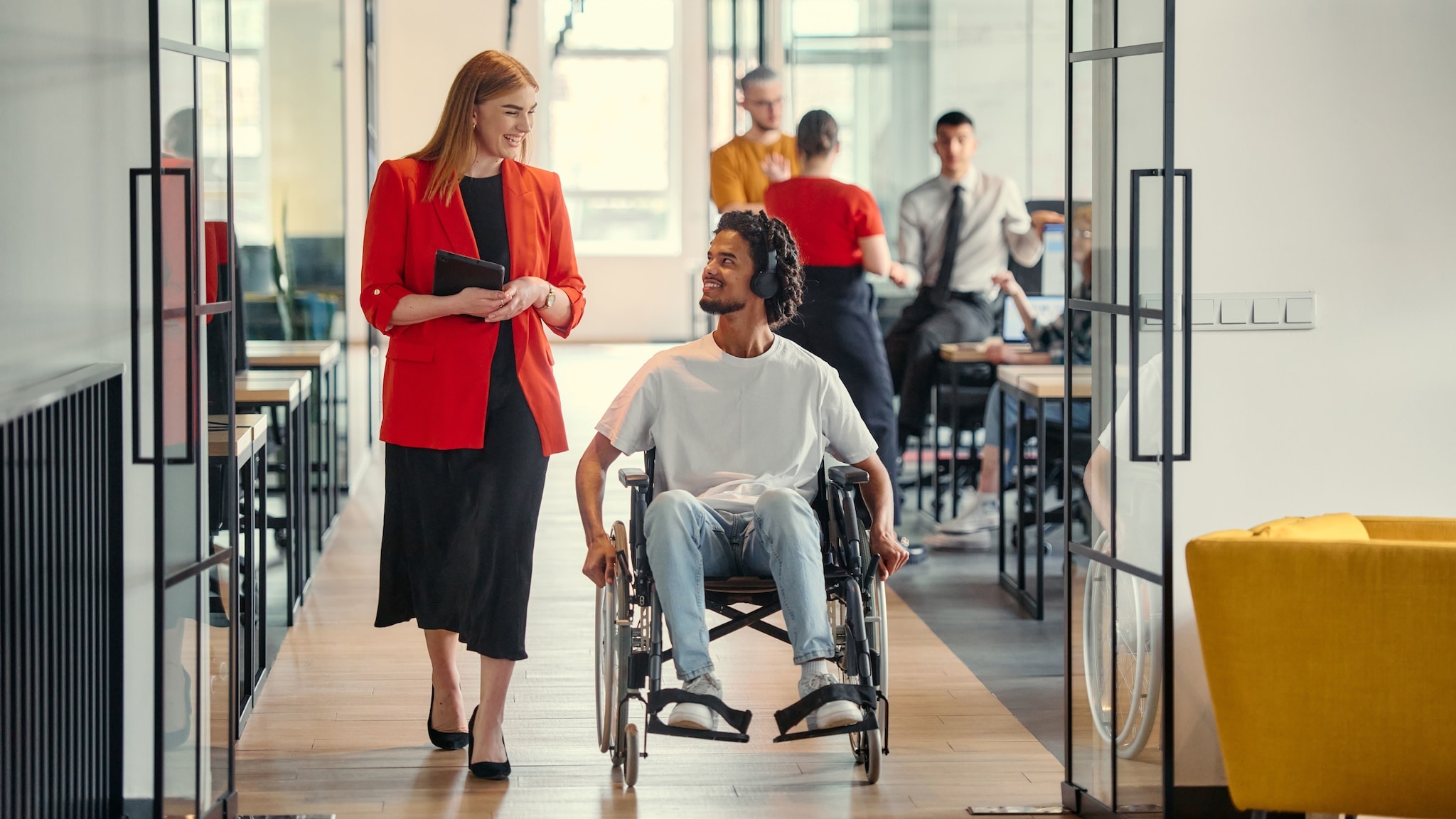 Eine Frau im roten Blazer und ein Rollstuhlfahrer mit Kopfhörern kommunizieren im Durchgang eines Großraumbüros. Eine Frau im roten Blazer und ein Rollstuhlfahrer mit Kopfhörern kommunizieren im Durchgang eines Großraumbüros.