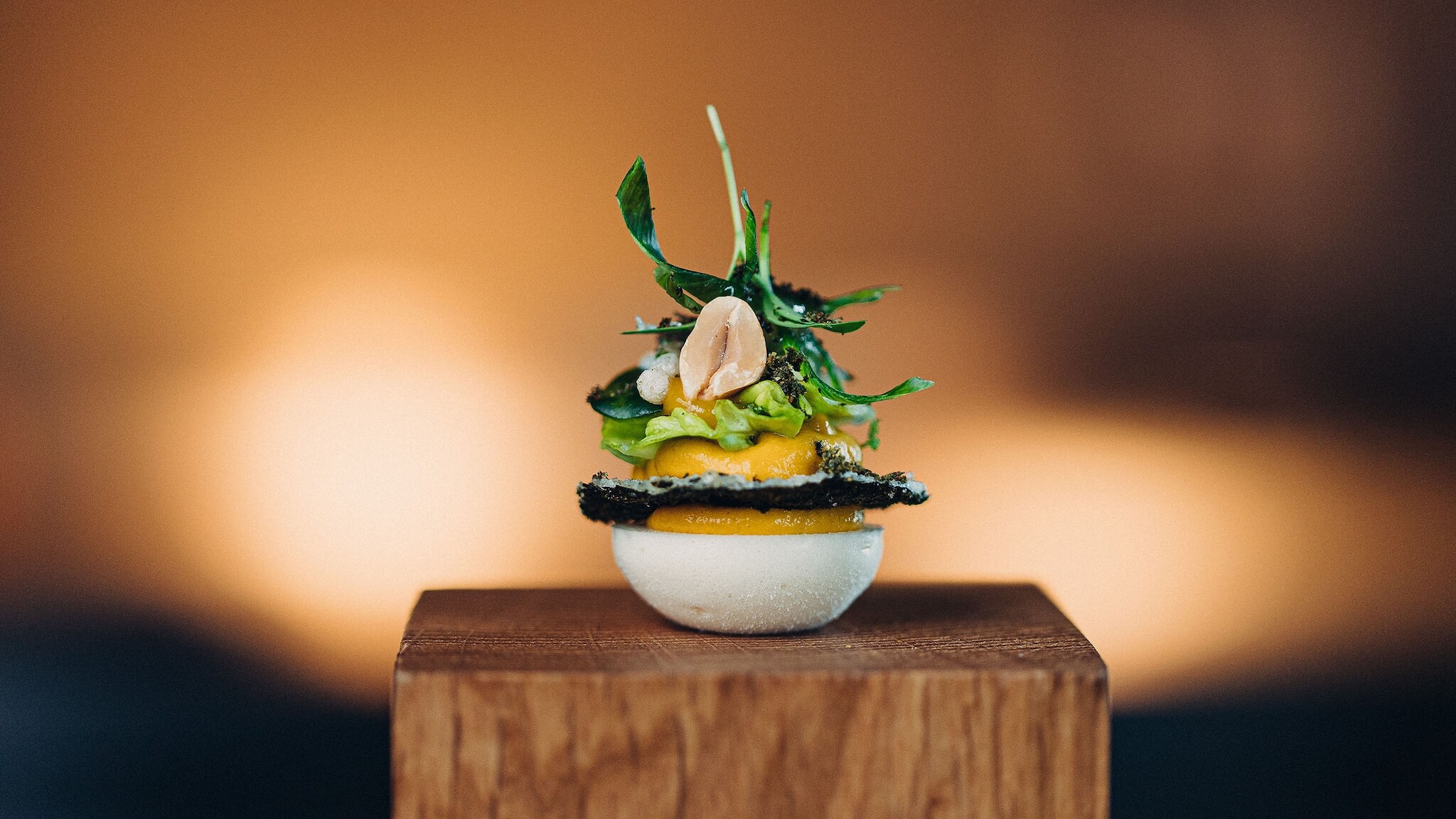 Eine exklusive Vorspeise aus geschichteten Zutaten, angerichtet auf einem Holzsockel vor unscharfen Hintergrund.