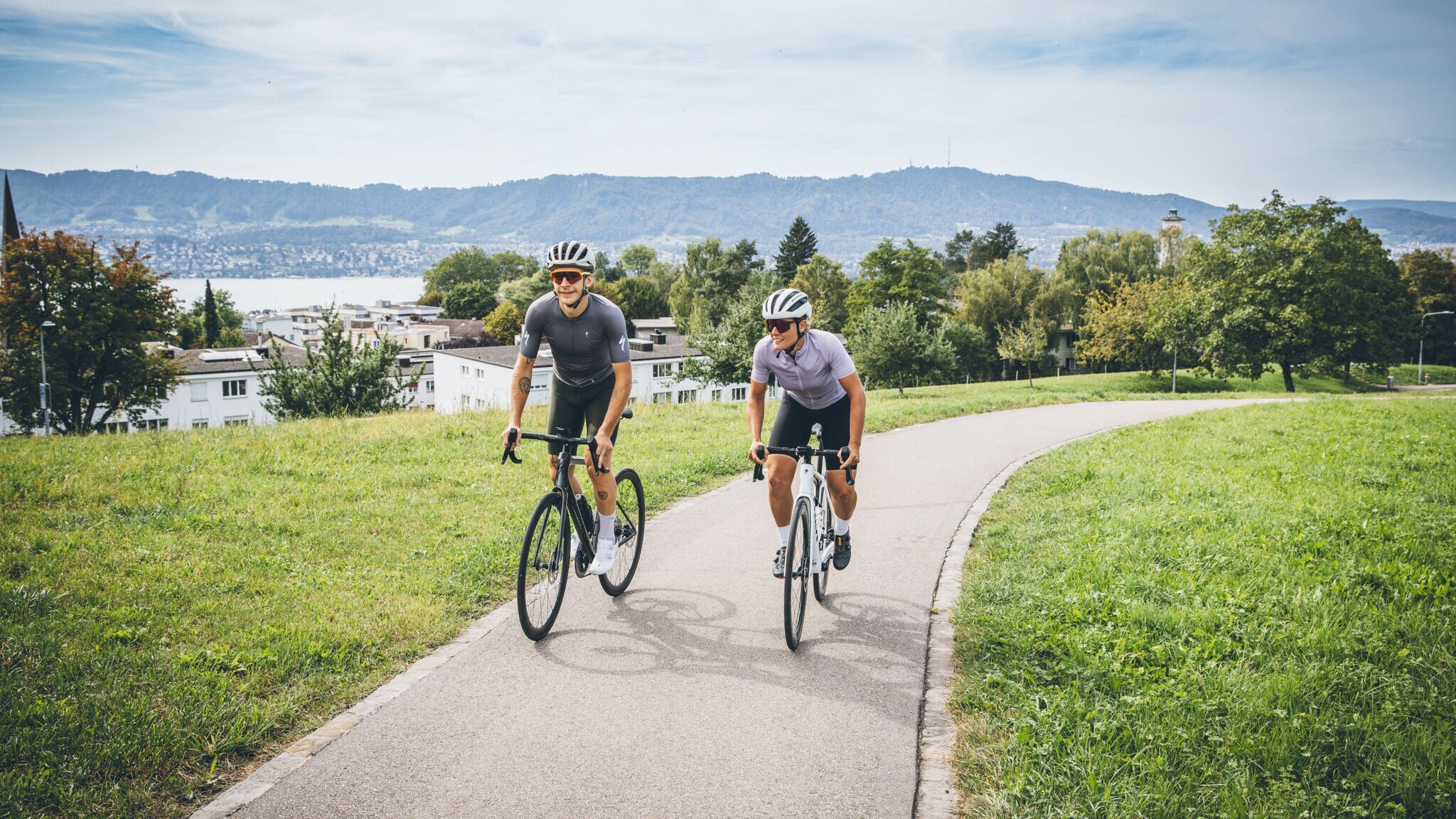 Zwei Personen auf Mountainbikes auf einem Radweg vor Seepanorama.