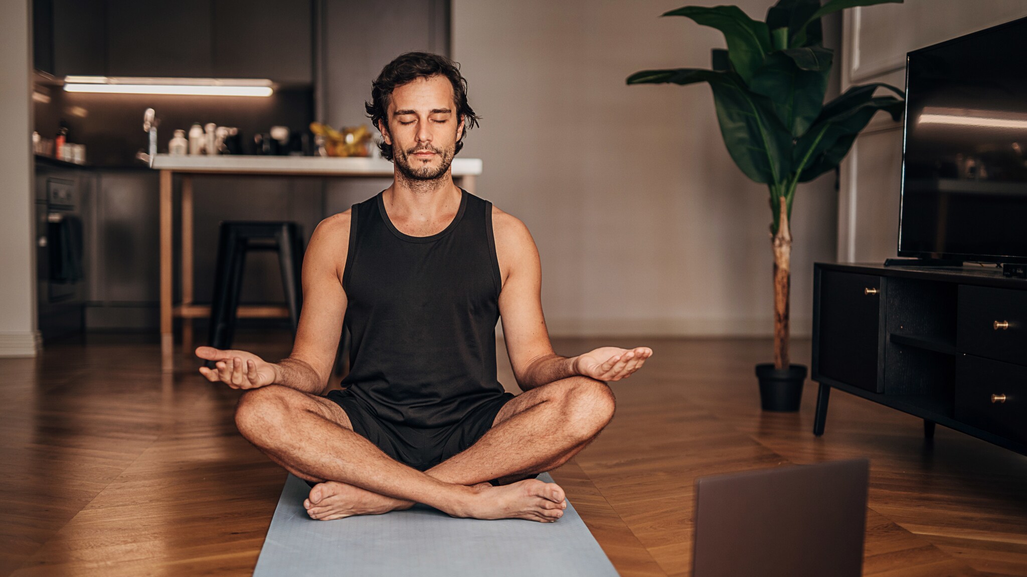 Ein Mann im schwarzen Sport-Outfit sitzt im Schneidersitz in Meditationspose mit geschlossenen Augen auf einer Yogamatte in einem Wohnzimmer.