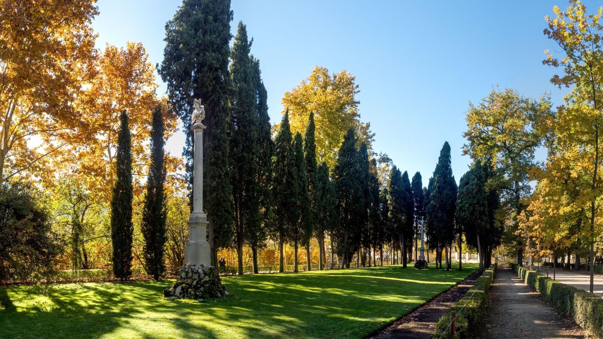 Idyllische Parkanlage mit teils herbstlich gefärbten Bäumen und Skulpturen auf Säulen unter blauem Himmel.