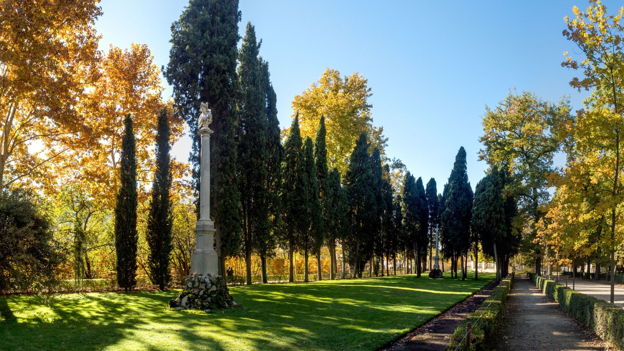 Idyllische Parkanlage mit teils herbstlich gefärbten Bäumen und Skulpturen auf Säulen unter blauem Himmel. Idyllische Parkanlage mit teils herbstlich gefärbten Bäumen und Skulpturen auf Säulen unter blauem Himmel.