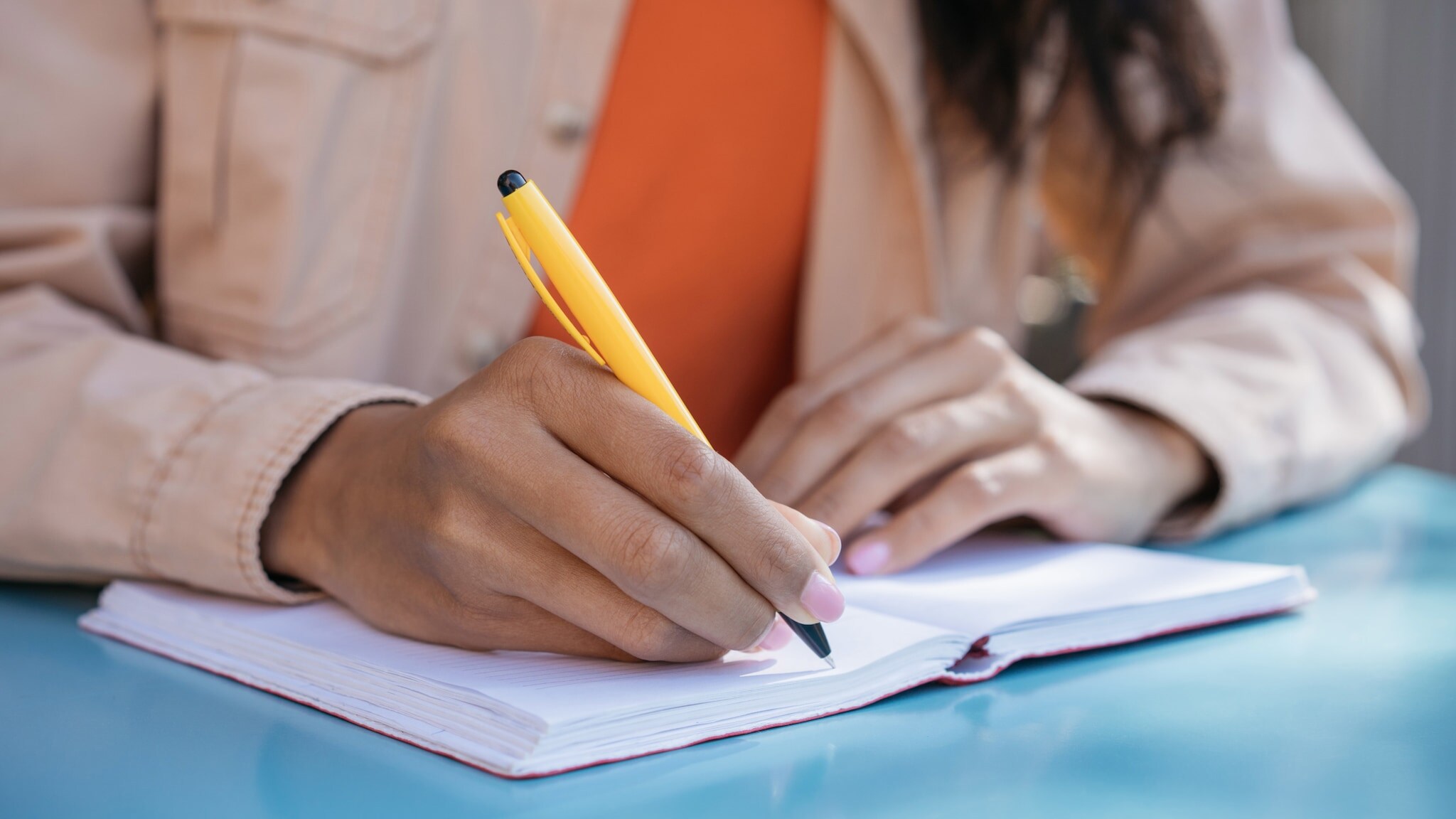 Nahaufnahme von zwei Händen über einem geöffneten Notizheft auf einem Tisch, in einer Hand ein gelber Kugelschreiber. Nahaufnahme von zwei Händen über einem geöffneten Notizheft auf einem Tisch, in einer Hand ein gelber Kugelschreiber.