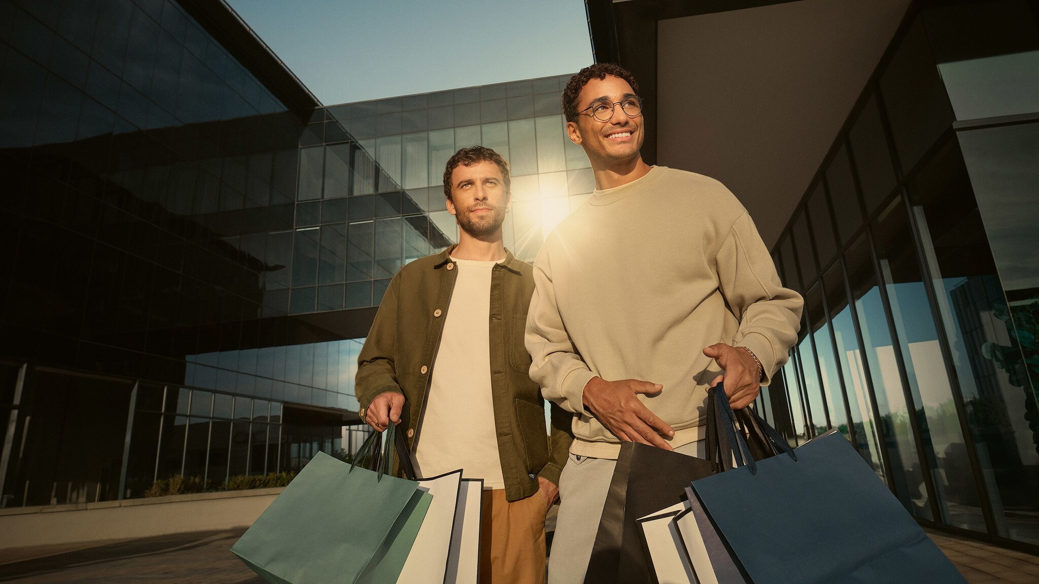Zwei moderne Männer mit mehreren Einkaufstüten in den Händen stehen vor einem modernen Gebäude mit Spiegelfassade.