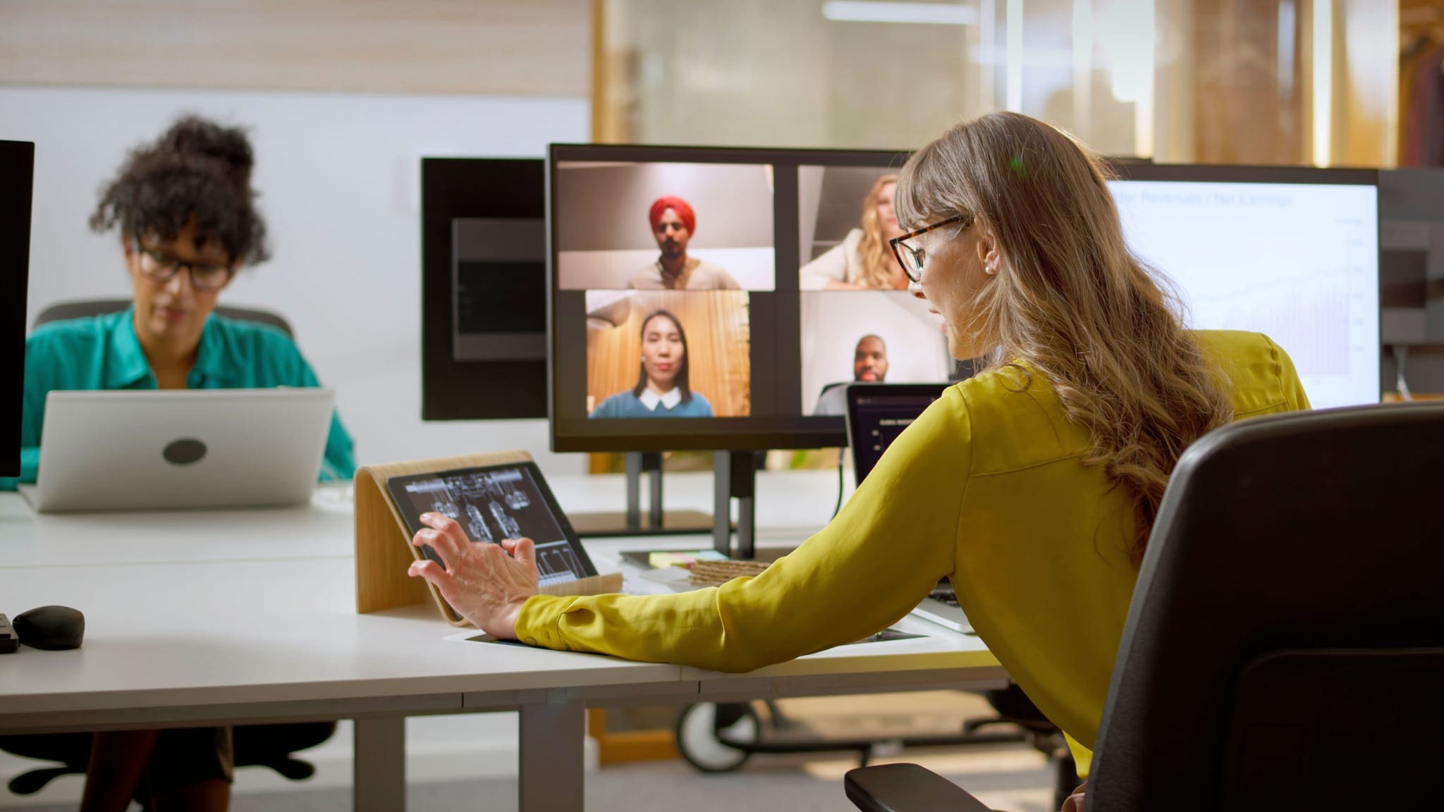 Zwei Frauen sitzen an Laptops an Schreibtischen. Auf einem Bildschirm sind Videocall-Kacheln weiterer Personen zu sehen. Zwei Frauen sitzen an Laptops an Schreibtischen. Auf einem Bildschirm sind Videocall-Kacheln weiterer Personen zu sehen.