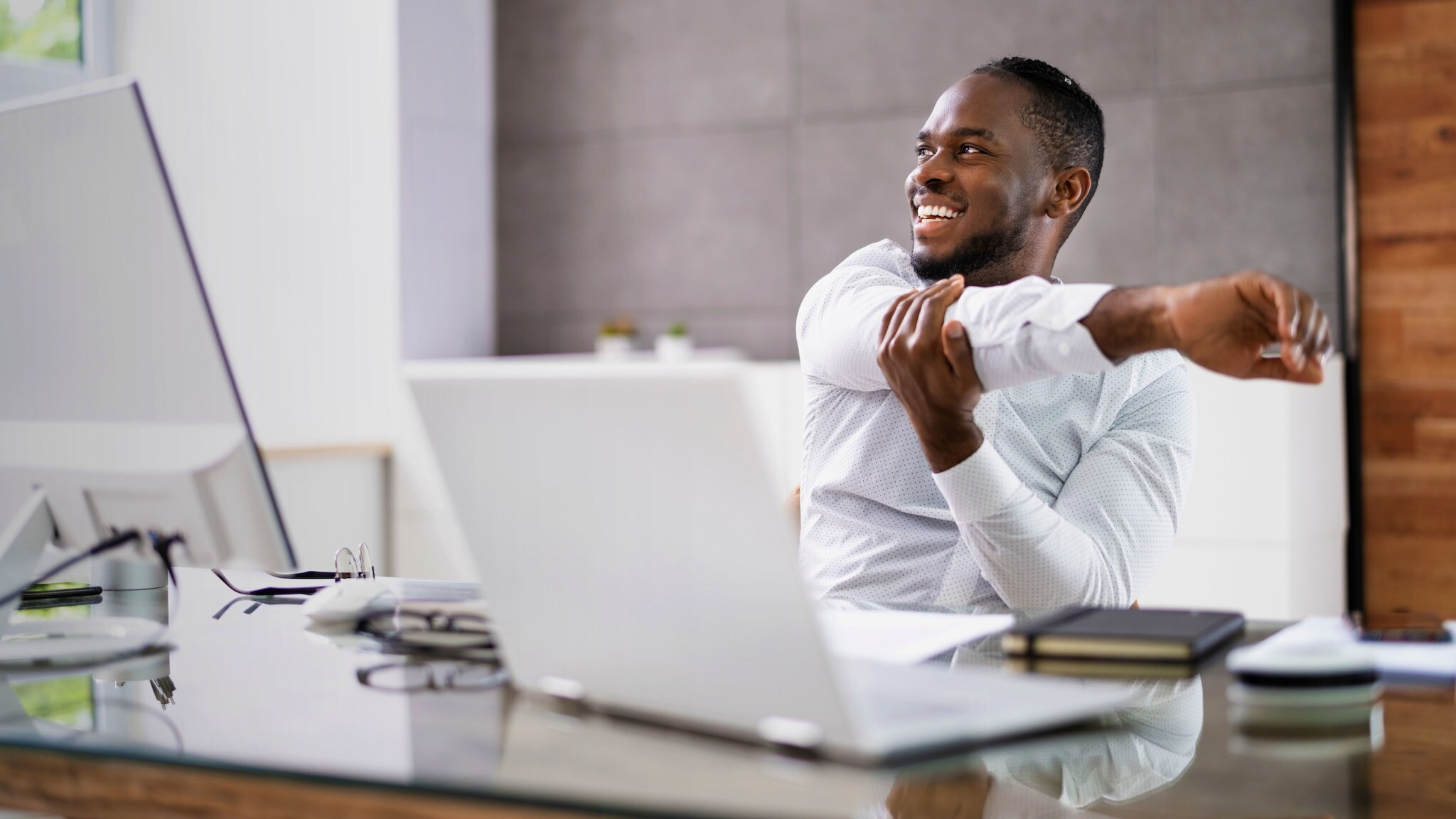 Ein Mann im weißen Hemd sitzt an einem Schreibtisch, lächelt und dehnt seinen Arm.