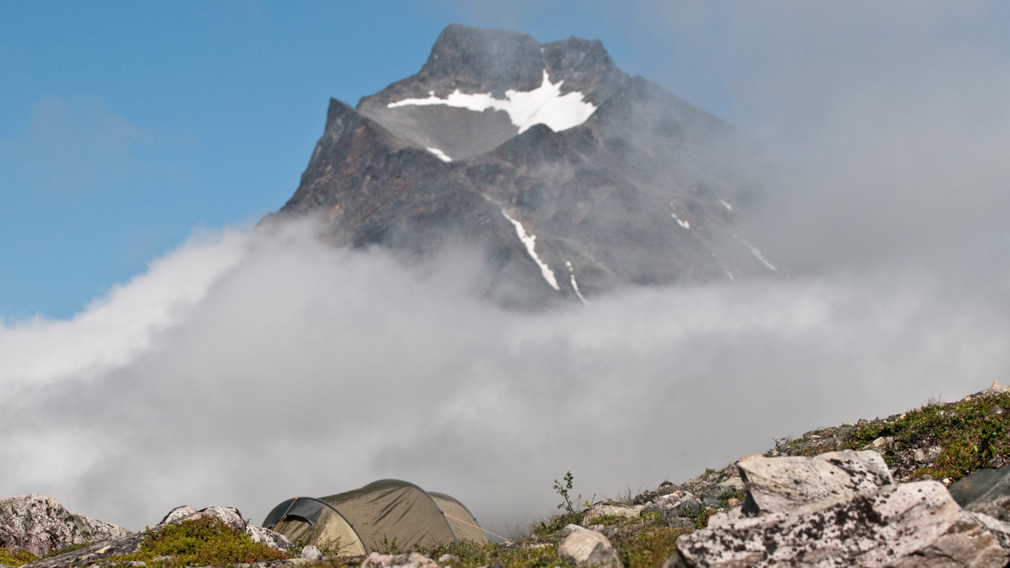 Grünes Zelt im Vordergrund, im Hintergrund ein schneebedeckter Berg mit Wolken und Felsen.