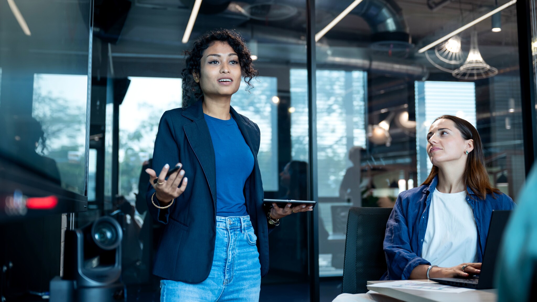 Eine Frau mit Stift und Tablet in der Hand spricht zu weiteren Personen in einem Raum mit gläsernen Wänden.