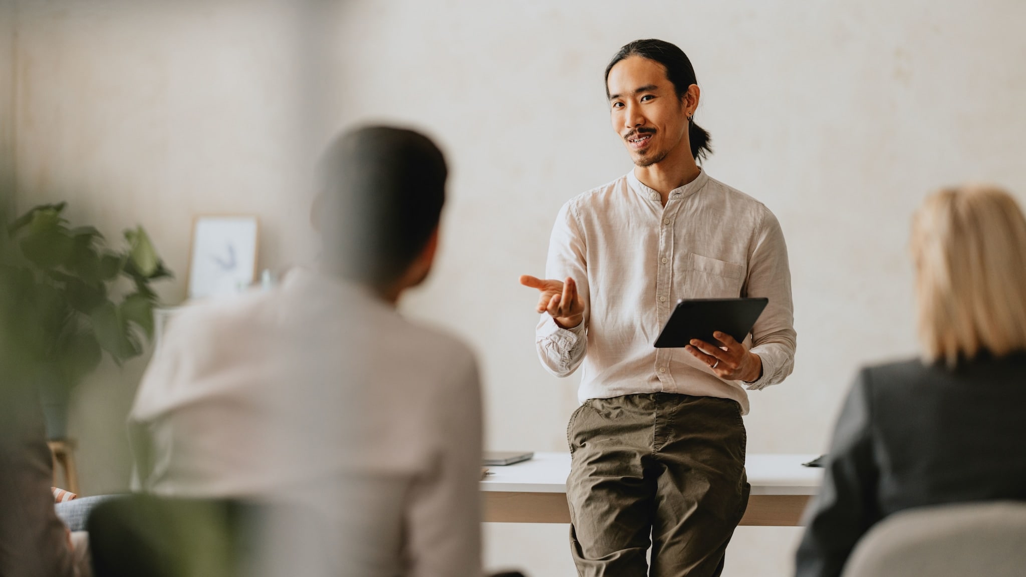 Ein Mann mit Tablet in der Hand lehnt an einem Tisch und kommuniziert mit sitzenden Personen. Ein Mann mit Tablet in der Hand lehnt an einem Tisch und kommuniziert mit sitzenden Personen.