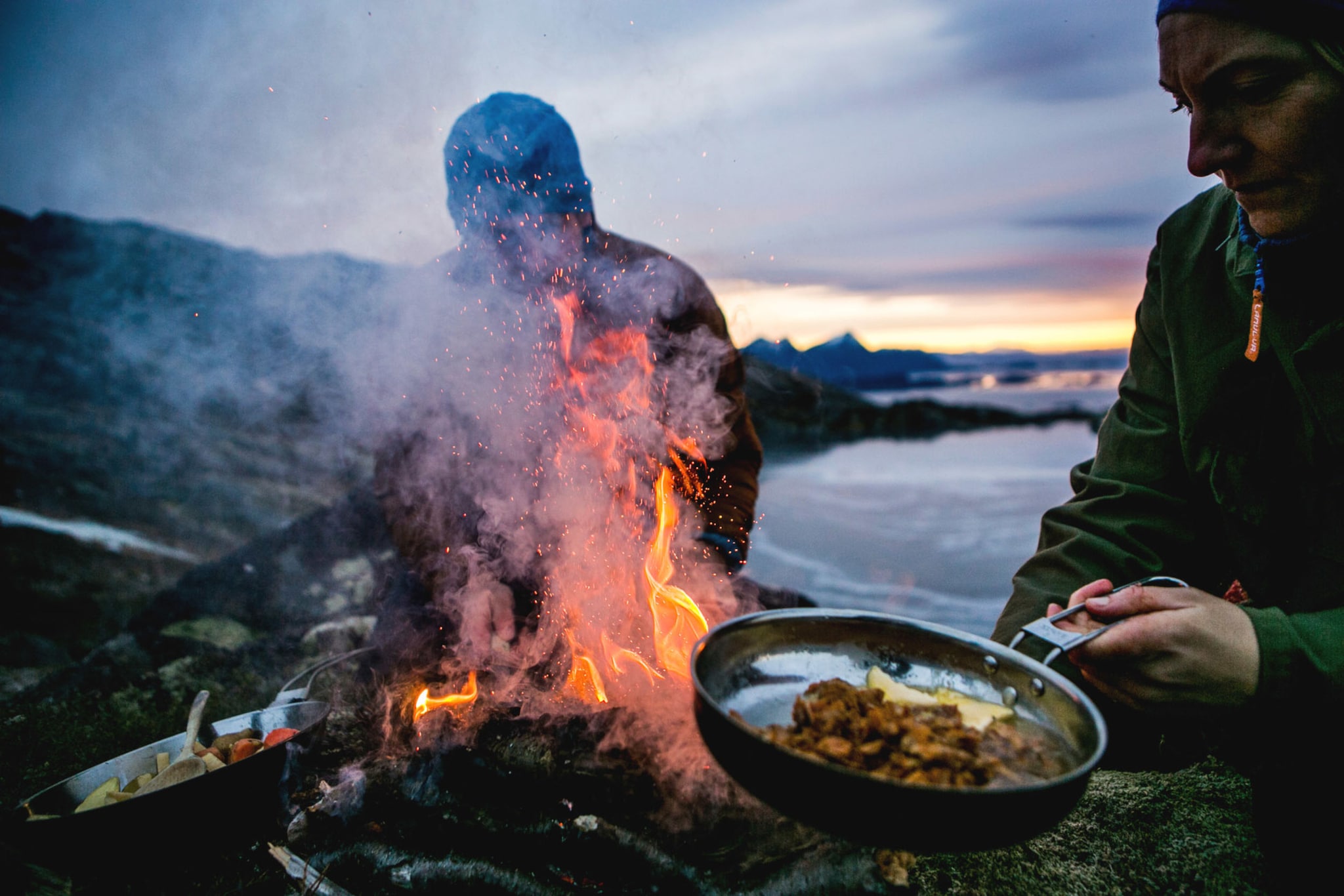 Zwei Personen bereiten Speisen in Pfannen über einem Feuer an einer schroffen Felsküste zu bei Abenddämmerung.