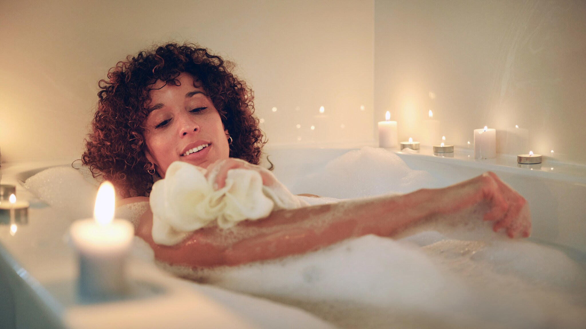 Eine Frau seift sich mit einem Schwamm ein in einer Badewanne voll Badeschaum mit Kerzen am Rand.