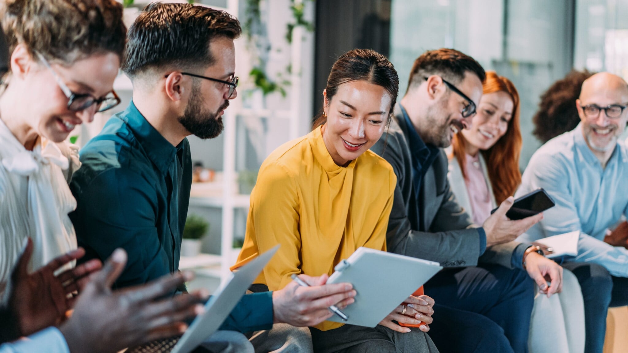 Mehrere Personen mit Notizheften und mobilen Endgeräten in den Händen sitzen gut gelaunt nebeneinander in einem Büro und unterhalten sich. Mehrere Personen mit Notizheften und mobilen Endgeräten in den Händen sitzen gut gelaunt nebeneinander in einem Büro und unterhalten sich.