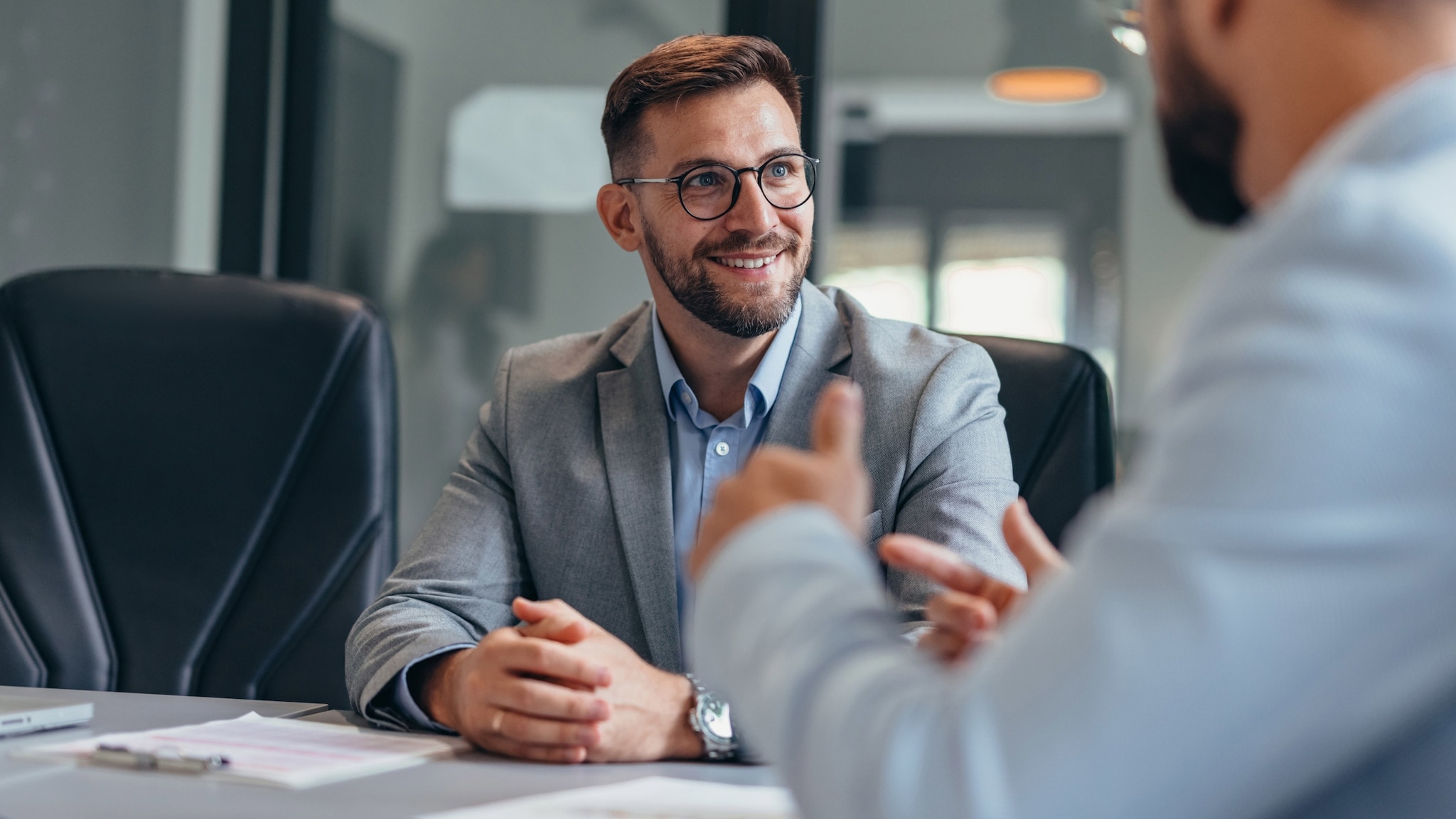 Ein Mann in Business-Kleidung sitzt einem gestikulierenden Mann an einem Konferenztisch lächelnd gegenüber. Ein Mann in Business-Kleidung sitzt einem gestikulierenden Mann an einem Konferenztisch lächelnd gegenüber.