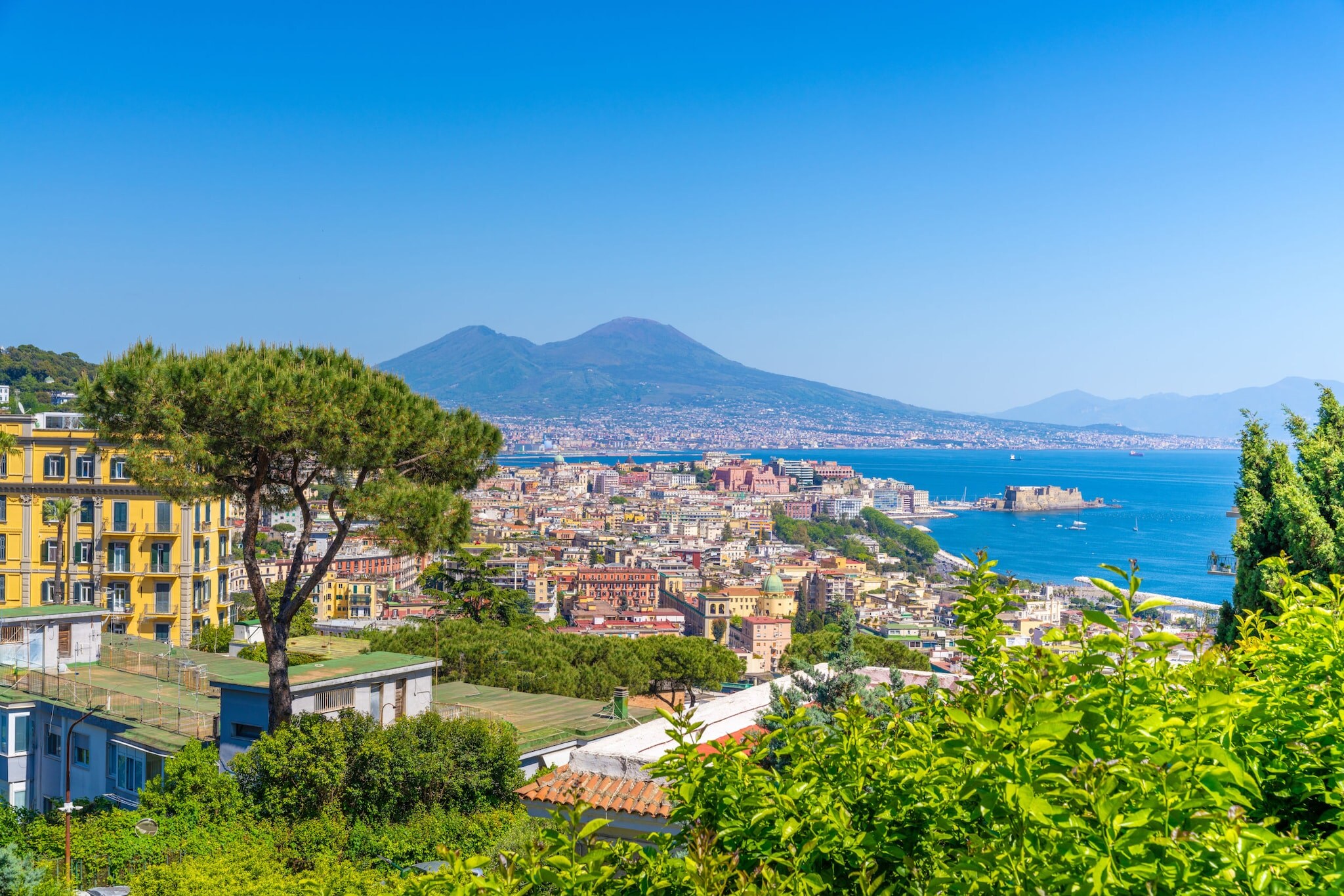 Blick von oben auf die Häuser von Neapel, im Hintergrund Meer und ein Vulkan.