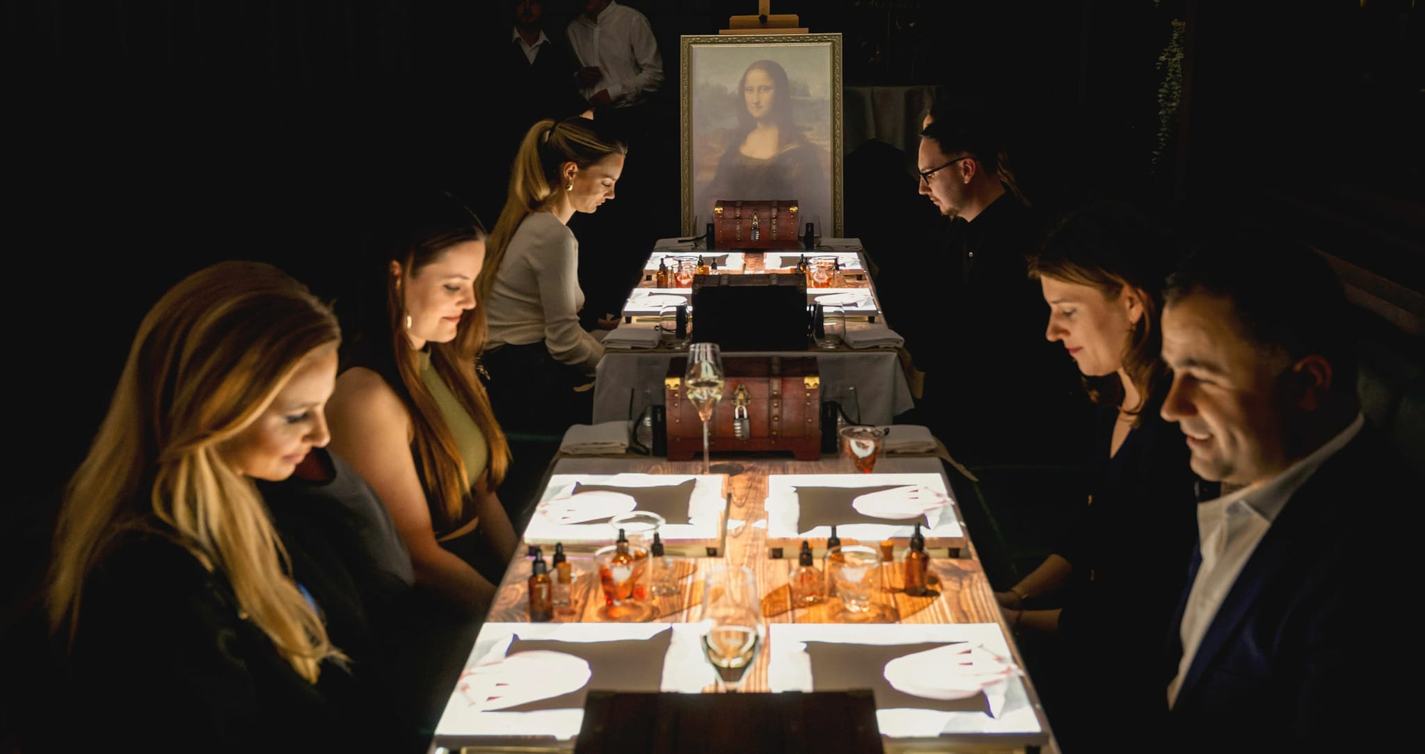 Mehrere Personen sitzen an Esstischen mit Lichtprojektionen neben einem Gemälde der Mona Lisa in einem dunklen Raum.