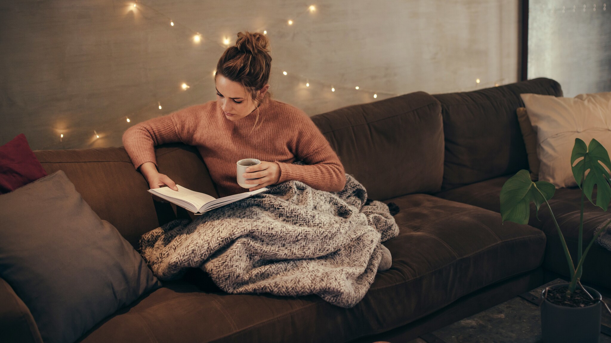 Eine Frau hockt auf einem Sofa mit einer Decke über den Beinen. In der einen Hand hat sie einen Becher, in der anderen ein aufgeschlagenes Buch, auf das sie schaut. Eine Frau hockt auf einem Sofa mit einer Decke über den Beinen. In der einen Hand hat sie einen Becher, in der anderen ein aufgeschlagenes Buch, auf das sie schaut.