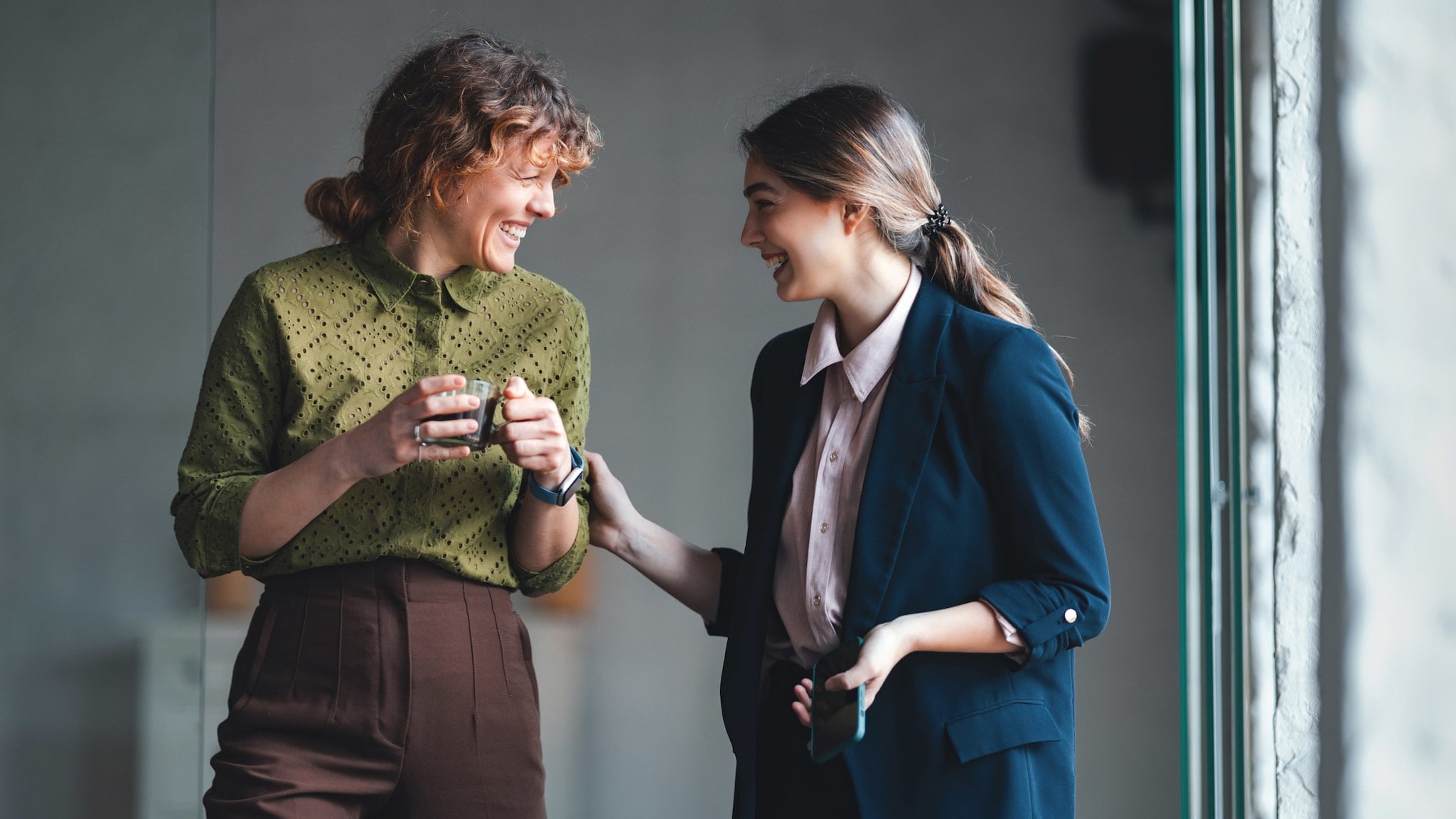 Zwei Frauen lächeln sich an, eine fasst der anderen, die eine Kaffeetasse in beiden Händen hält, dabei behutsam an den Arm. Zwei Frauen lächeln sich an, eine fasst der anderen, die eine Kaffeetasse in beiden Händen hält, dabei behutsam an den Arm.
