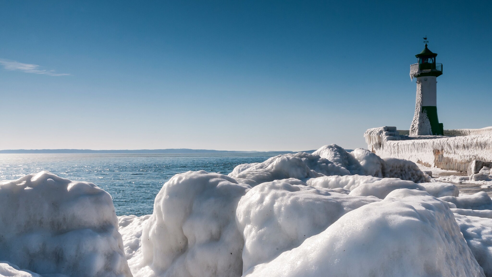 Winterlicher Küstenabschnitt mit Eisblöcken vor einem Leuchtturm unter blauem Himmel bei Sonnenschein.