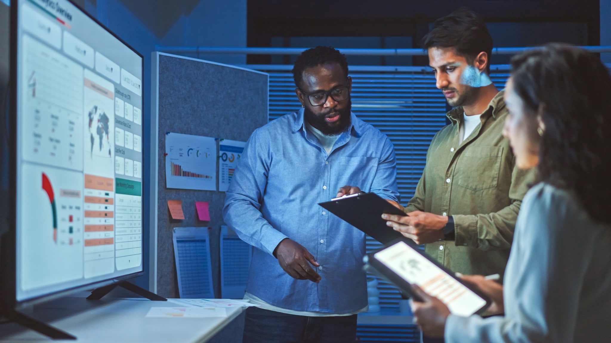 Drei Personen stehen mit Arbeitsunterlagen vor einem digitalen Dashboard in einem Konferenzraum.