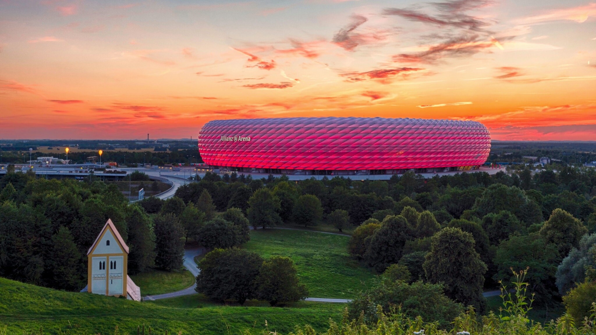 Panoramaaufnahme eines pink leuchtenden Fußballstadions unter orange gefärbtem Himmel bei Sonnenuntergang, im Vordergrund Wiesenfläche mit Wald und Kapelle.