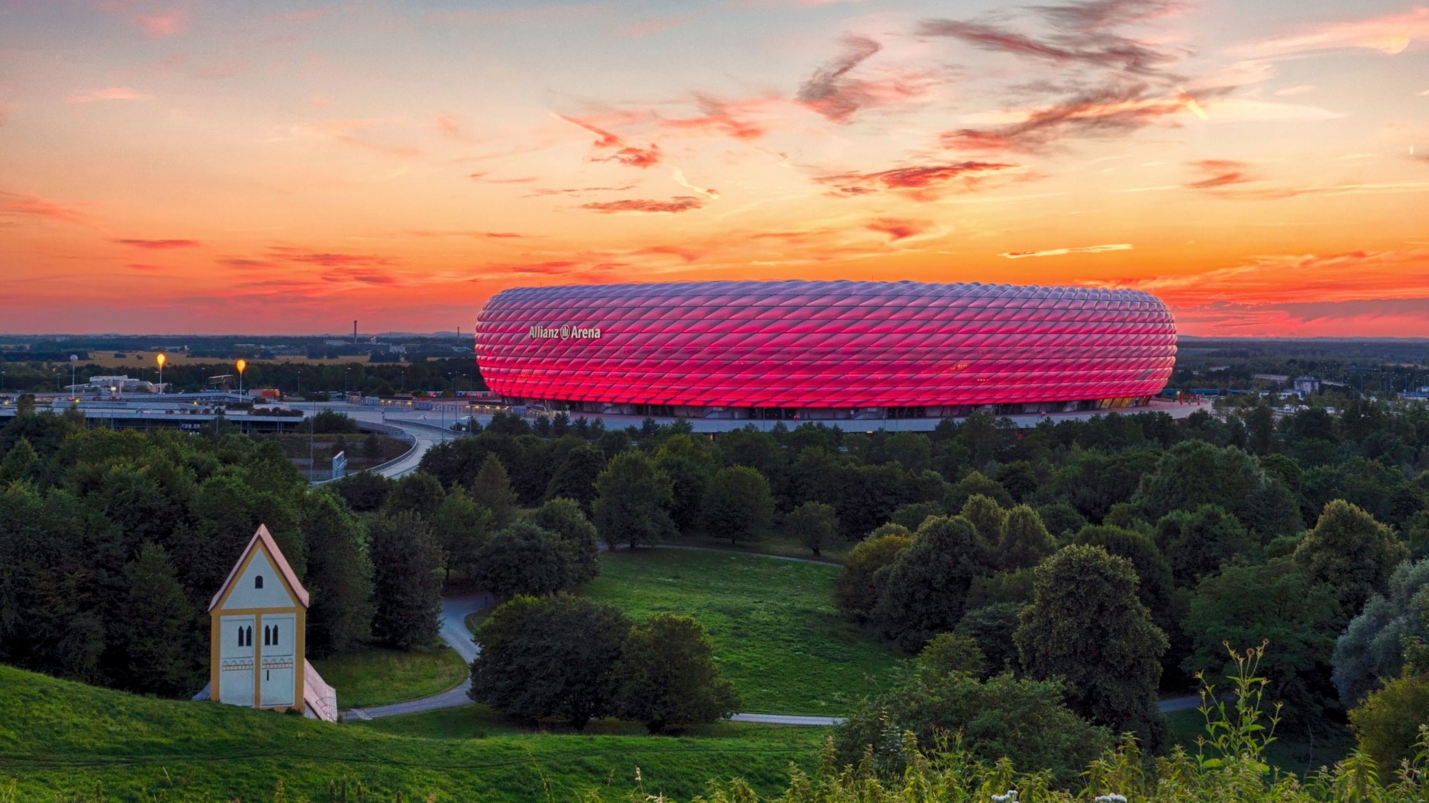 Panoramaaufnahme eines pink leuchtenden Fußballstadions unter orange gefärbtem Himmel bei Sonnenuntergang, im Vordergrund Wiesenfläche mit Wald und Kapelle. Panoramaaufnahme eines pink leuchtenden Fußballstadions unter orange gefärbtem Himmel bei Sonnenuntergang, im Vordergrund Wiesenfläche mit Wald und Kapelle.
