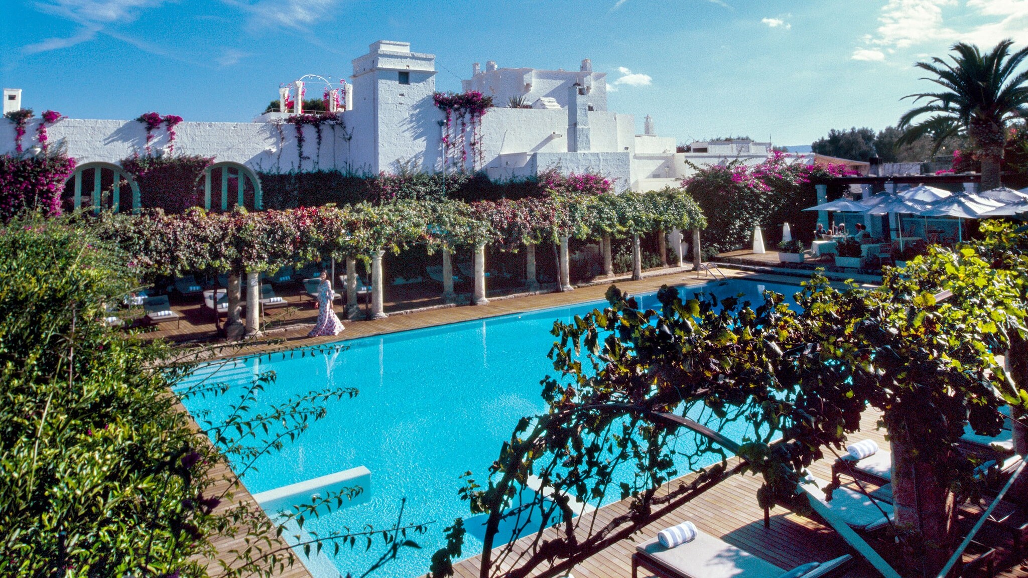 Idyllische, mit Weinranken begrünte Poolanlage in einem Hotelresort mit weißen Häusern unter blauem Himmel.
