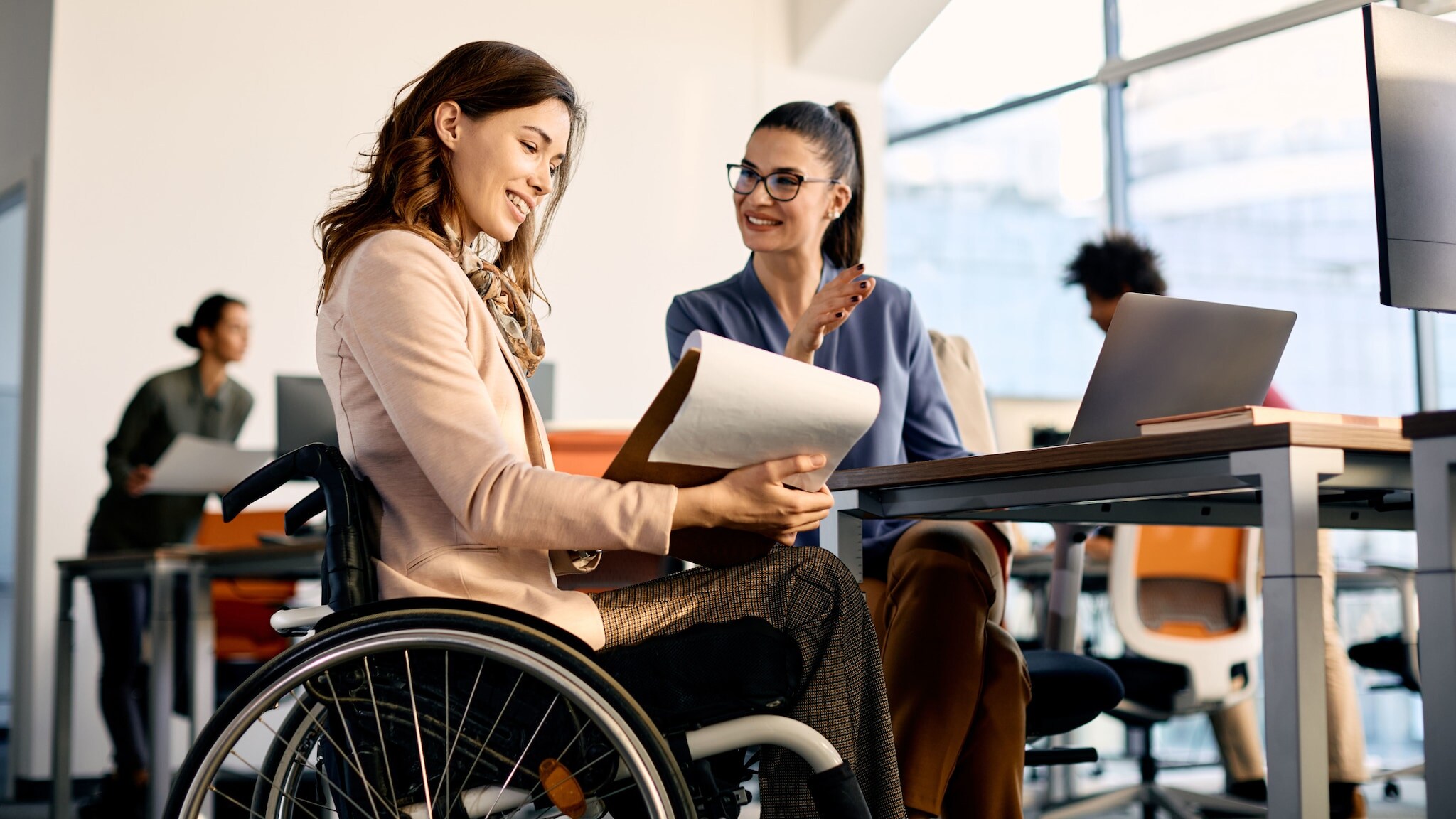 Eine Frau im Rollstuhl mit Klemmbrett in der Hand sitzt an einem Tisch neben einer weiteren Frau in einem hellen Raum.