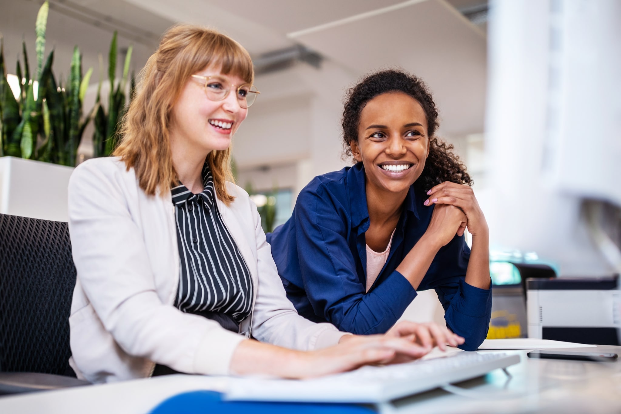 Zwei lächelnde Frauen, eine sitzt, die andere beugt sich vor und stützt die Ellenbogen auf den Tisch, schauen gemeinsam auf einen Computerbildschirm. Zwei lächelnde Frauen, eine sitzt, die andere beugt sich vor und stützt die Ellenbogen auf den Tisch, schauen gemeinsam auf einen Computerbildschirm.