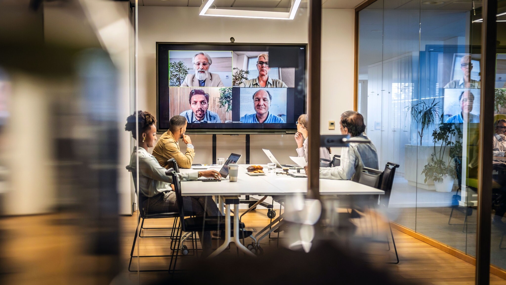 Vier Personen sitzen an einem Konferenztisch vor einem großen Bildschirm mit Gesichtern von vier weiteren Personen während eines Online-Meetings in einem Büro.