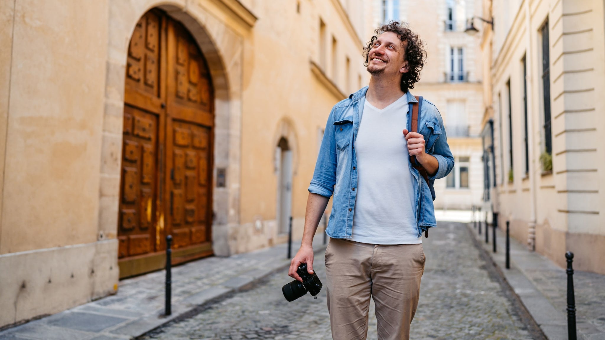 Ein Mann in legerer Kleidung geht mit einer Kamera in der Hand und Tasche über der Schulter durch eine schmale Straße und blickt lächelnd in die Höhe. Ein Mann in legerer Kleidung geht mit einer Kamera in der Hand und Tasche über der Schulter durch eine schmale Straße und blickt lächelnd in die Höhe.