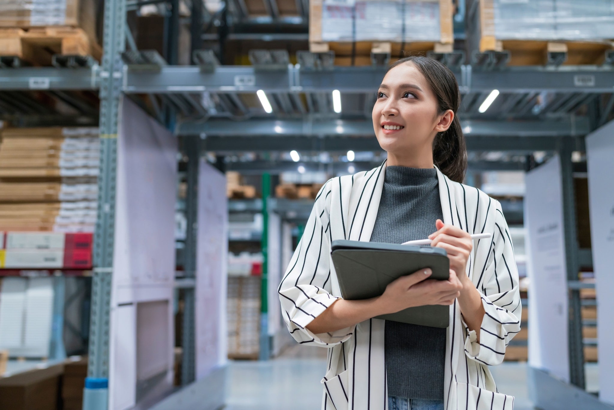 Eine lächelnde Frau steht mit Tablet und Stift in den Händen in einer Lagerhalle. Eine lächelnde Frau steht mit Tablet und Stift in den Händen in einer Lagerhalle.