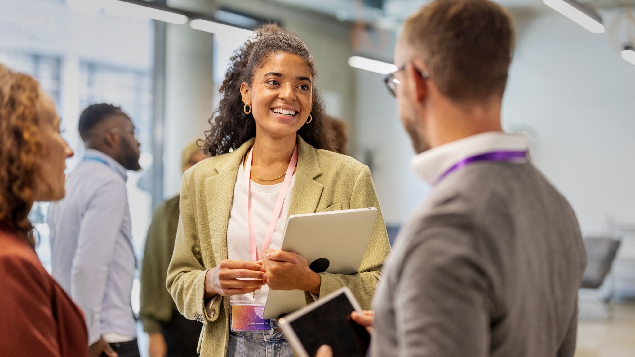Eine junge Frau im Blazer mit Laptop in der Hand steht in einem Konferenzraum und unterhält sich mit zwei weiteren Personen. Eine junge Frau im Blazer mit Laptop in der Hand steht in einem Konferenzraum und unterhält sich mit zwei weiteren Personen.