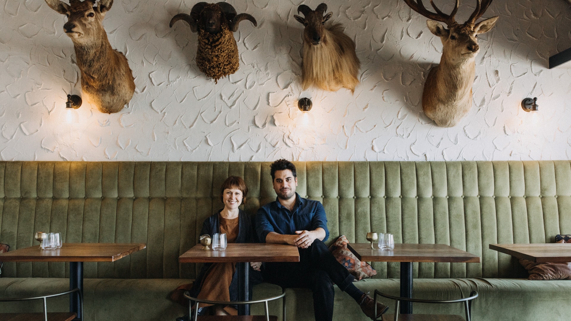 Eine Frau und ein Mann sitzen an einem Bistrotisch nebeneinander auf einer langen Bank aus grünen Samt an einer Wand unter vier ausgestopften Hirschköpfen.