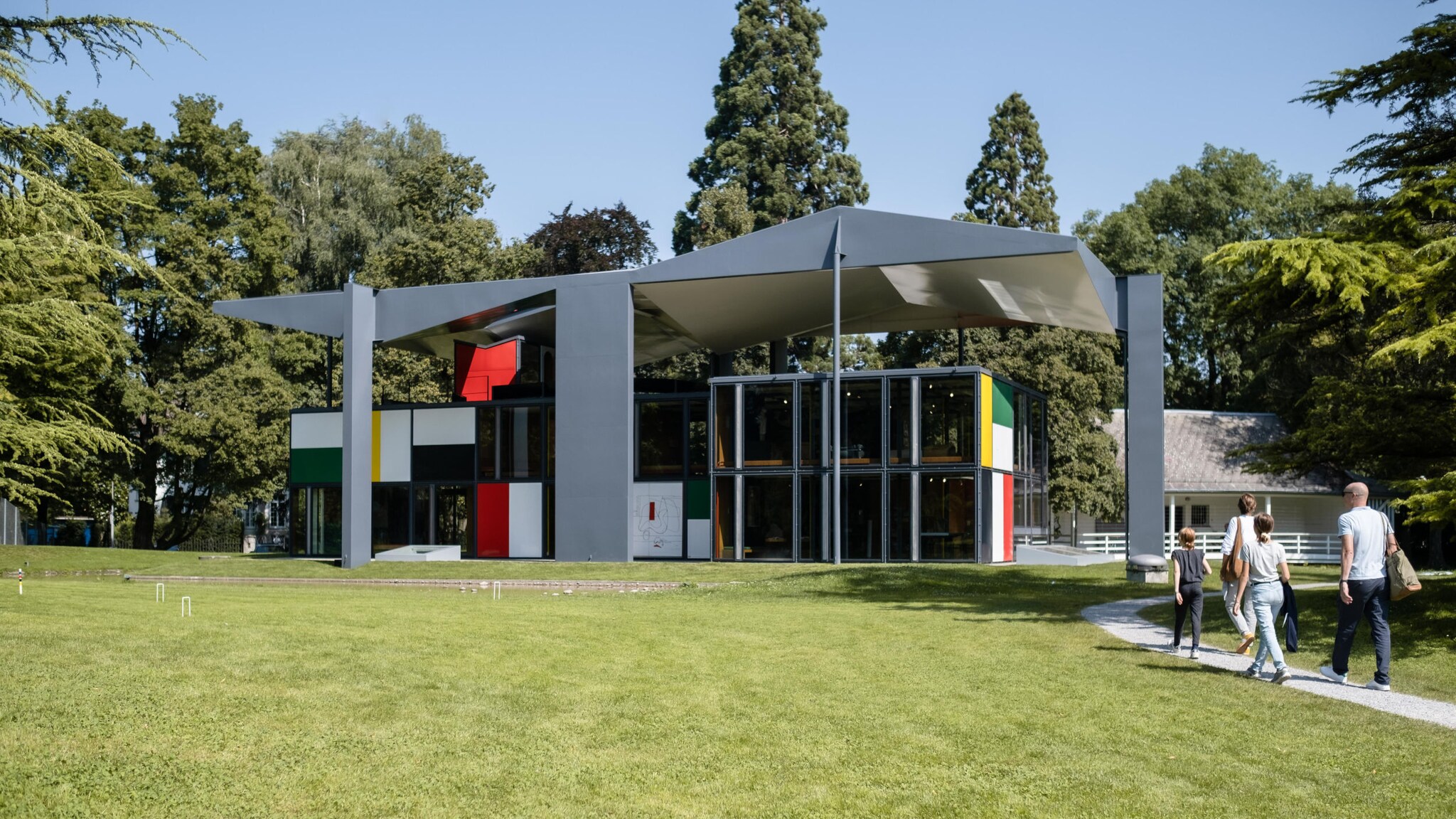 Eine Familie läuft auf ein modernes, fragmentiertes Gebäude im Le Corbusier-Stil in einer Parkanlage zu.