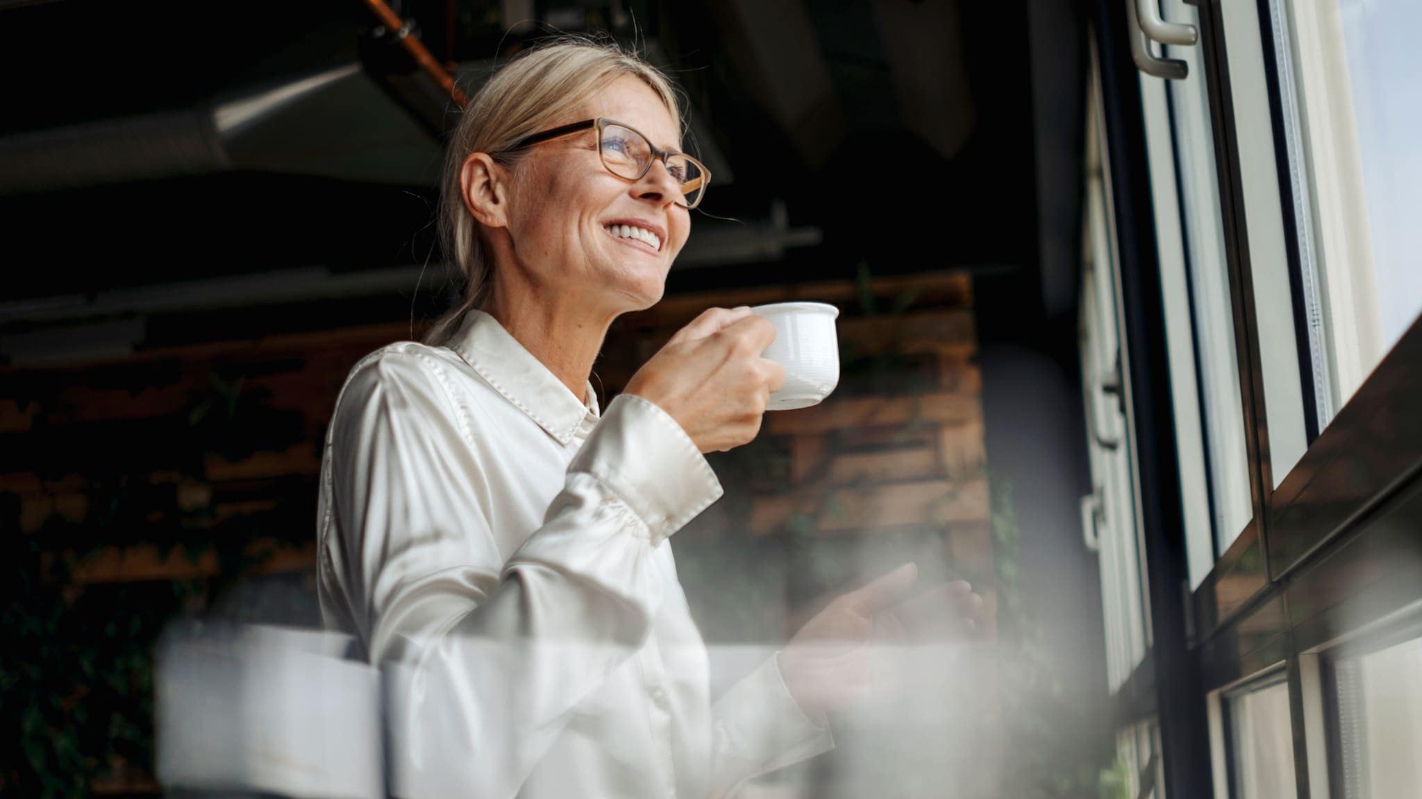 Eine lächelnde Frau in glänzender Bluse steht entspannt mit Kaffeetasse in der Hand an einem Fenster und blickt hinaus. Eine lächelnde Frau in glänzender Bluse steht entspannt mit Kaffeetasse in der Hand an einem Fenster und blickt hinaus.