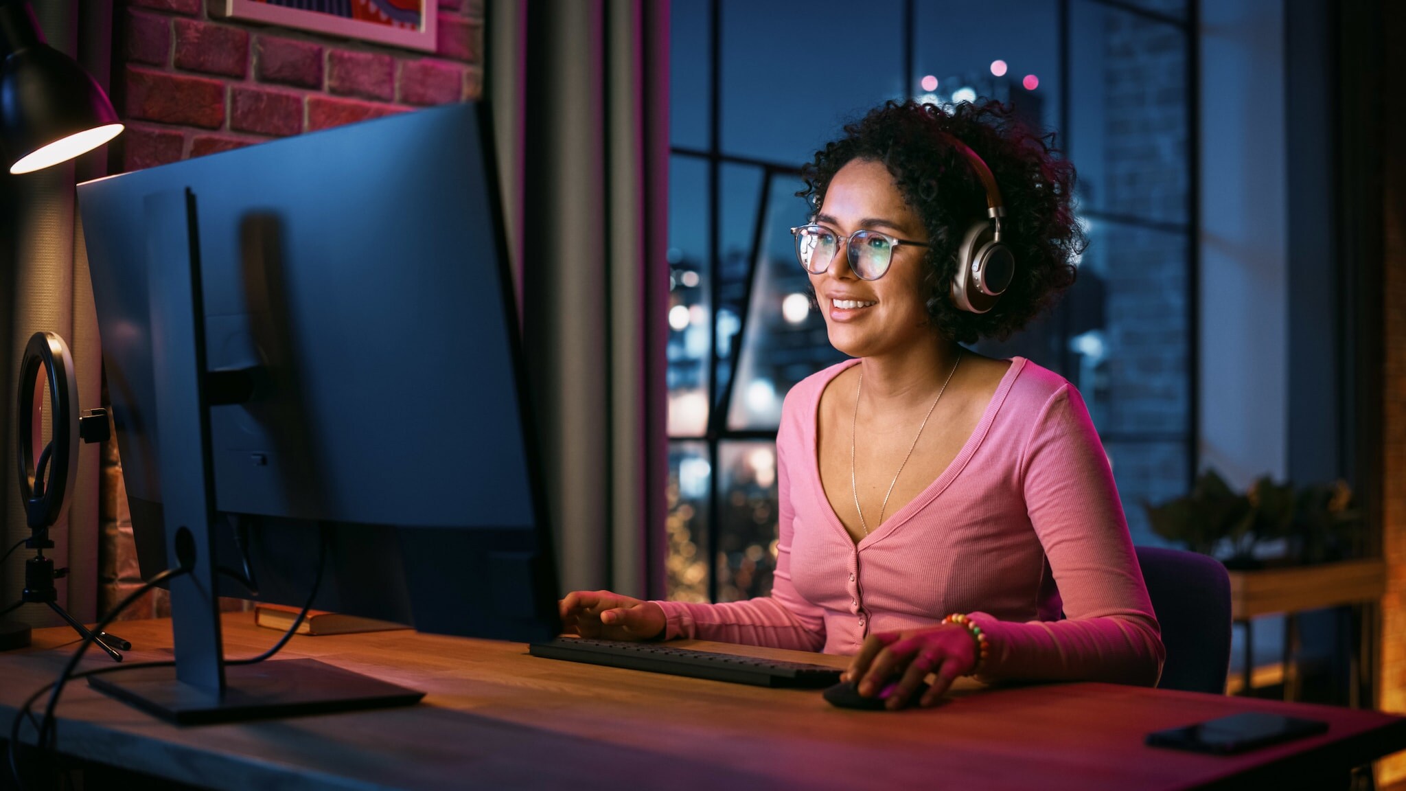 Eine lächelnde Frau im rosa Shirt sitzt mit Kopfhörern an einem Schreibtisch vor einem Computer in einem Raum in urbaner Umgebung bei Nacht.