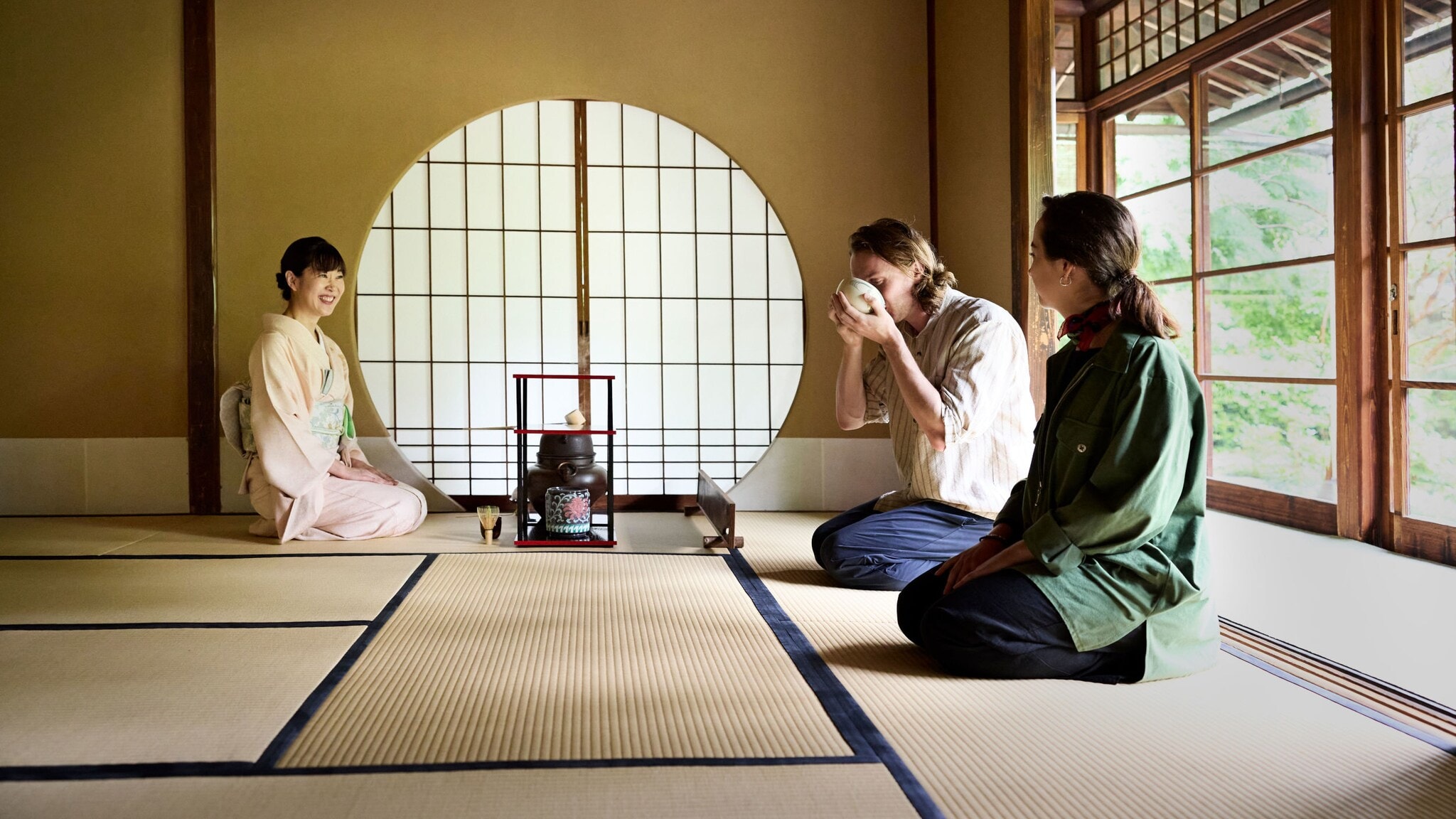 Eine Geisha sitzt zwei Personen, eine trinkt aus einer Schale, im Fersensitz gegenüber während einer Teezeremonie in einem hellen Raum im japanischen Stil mit Tatami-Matten und Gartenblick. Eine Geisha sitzt zwei Personen, eine trinkt aus einer Schale, im Fersensitz gegenüber während einer Teezeremonie in einem hellen Raum im japanischen Stil mit Tatami-Matten und Gartenblick.