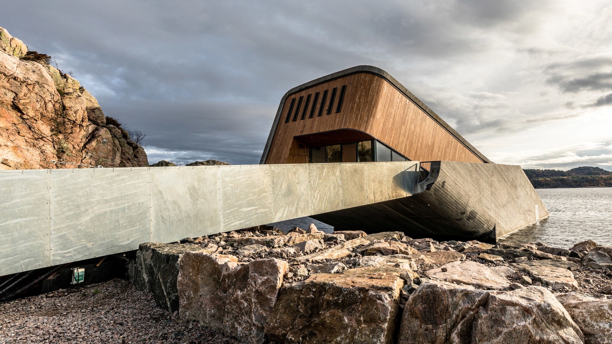 Eingangsbereich mit Holzfassade eines modernen Gebäudes, das schräg ins Wasser gebaut ist an einer Felsküste.