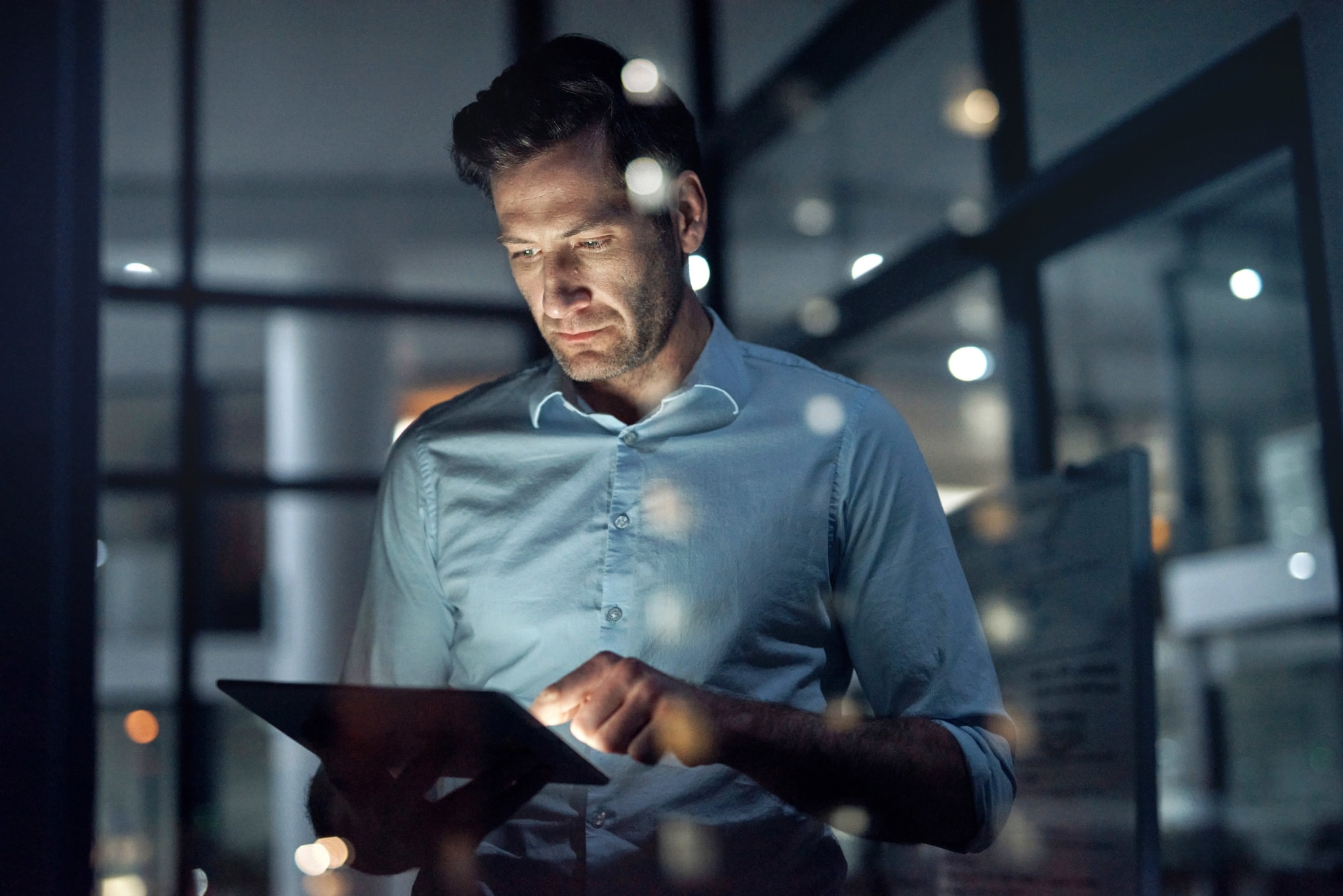 Ein Mann steht in einem dunklen Raum und hält ein Tablet in seinen Händen, dessen Displaylicht sein Gesicht anleuchtet. Ein Mann steht in einem dunklen Raum und hält ein Tablet in seinen Händen, dessen Displaylicht sein Gesicht anleuchtet.