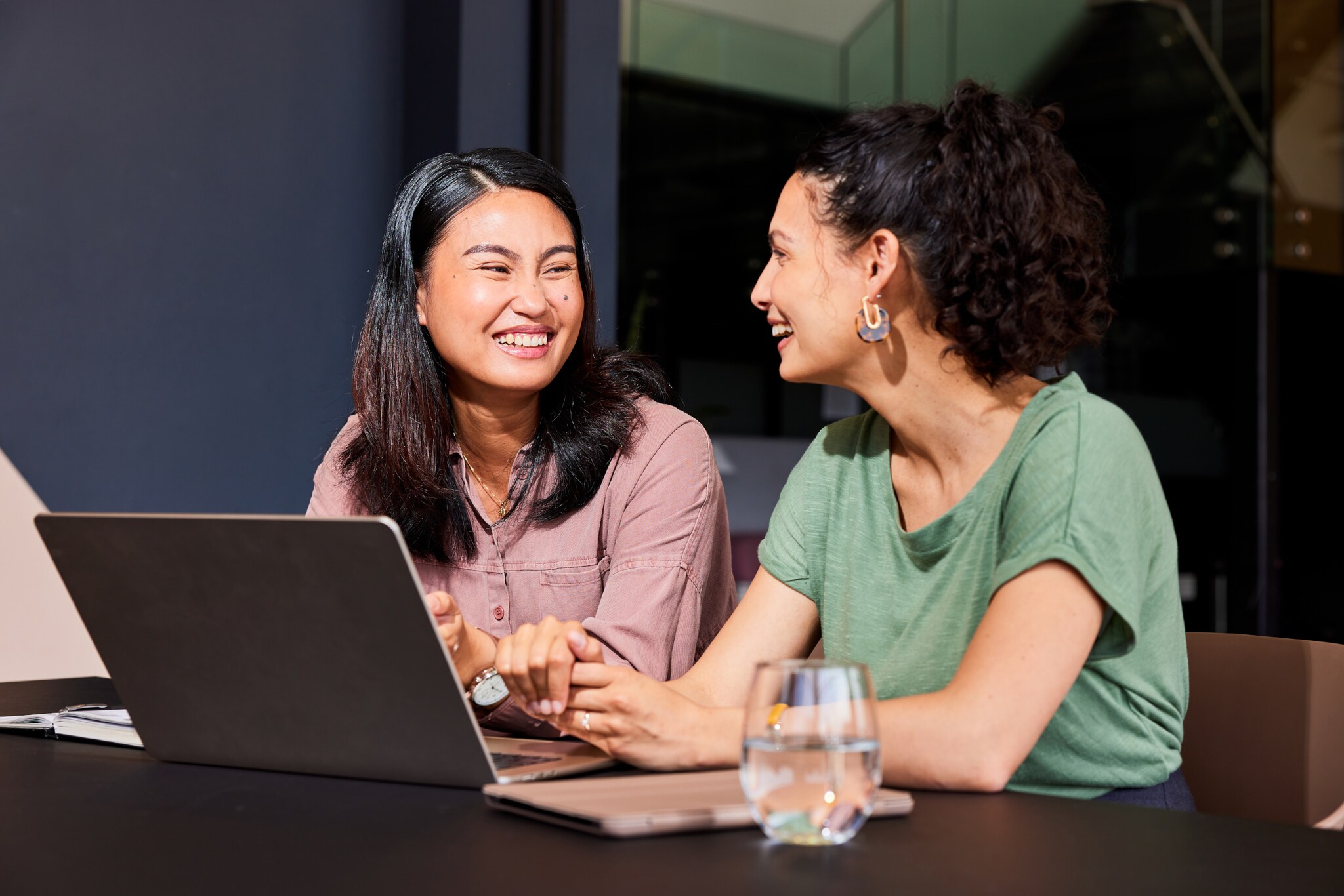 Zwei sich zugewandte lachende Frauen sitzen nebeneinander an einem Tisch, vor ihnen steht ein Laptop. Zwei sich zugewandte lachende Frauen sitzen nebeneinander an einem Tisch, vor ihnen steht ein Laptop.