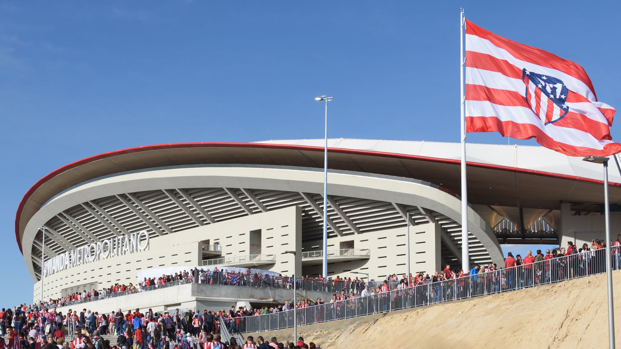Viele Personen vor dem Eingang eines Fußballstadions mit einer rot-weiß-gestreiften Flagge mit Wappen vor blauem Himmel.