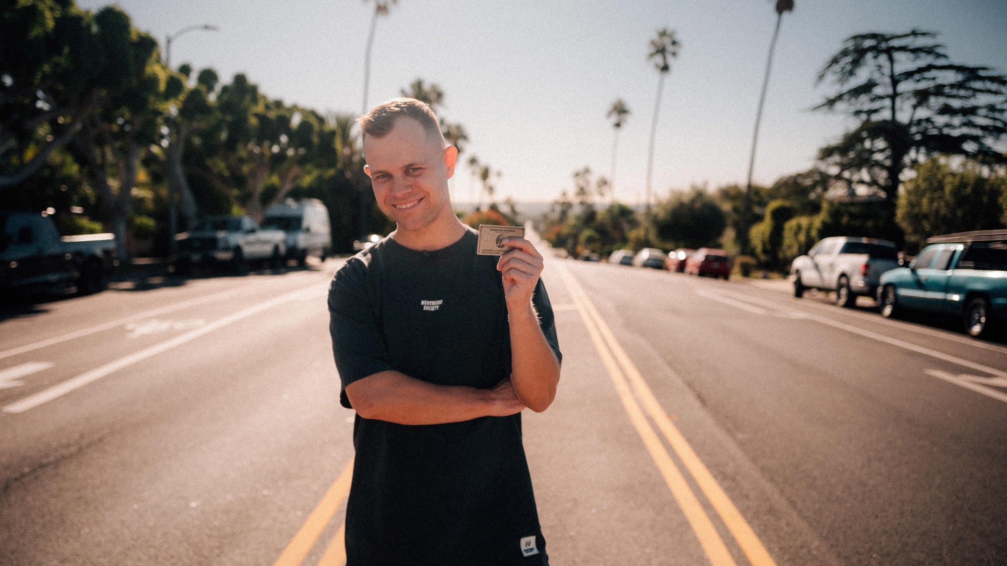 Benjamin Diedering posiert lächelnd mit einer Kreditkarte von American Express auf einer palmengesäumten Straße. Benjamin Diedering posiert lächelnd mit einer Kreditkarte von American Express auf einer palmengesäumten Straße.