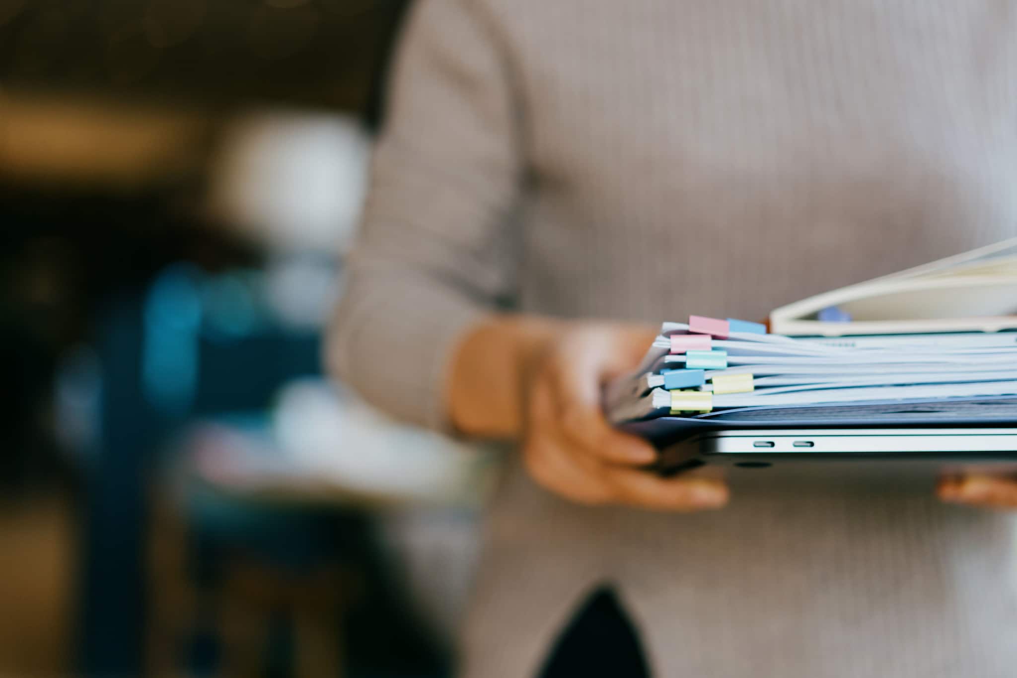 Eine Person trägt mit beiden Händen vor sich einen Laptop und Papierunterlagen.