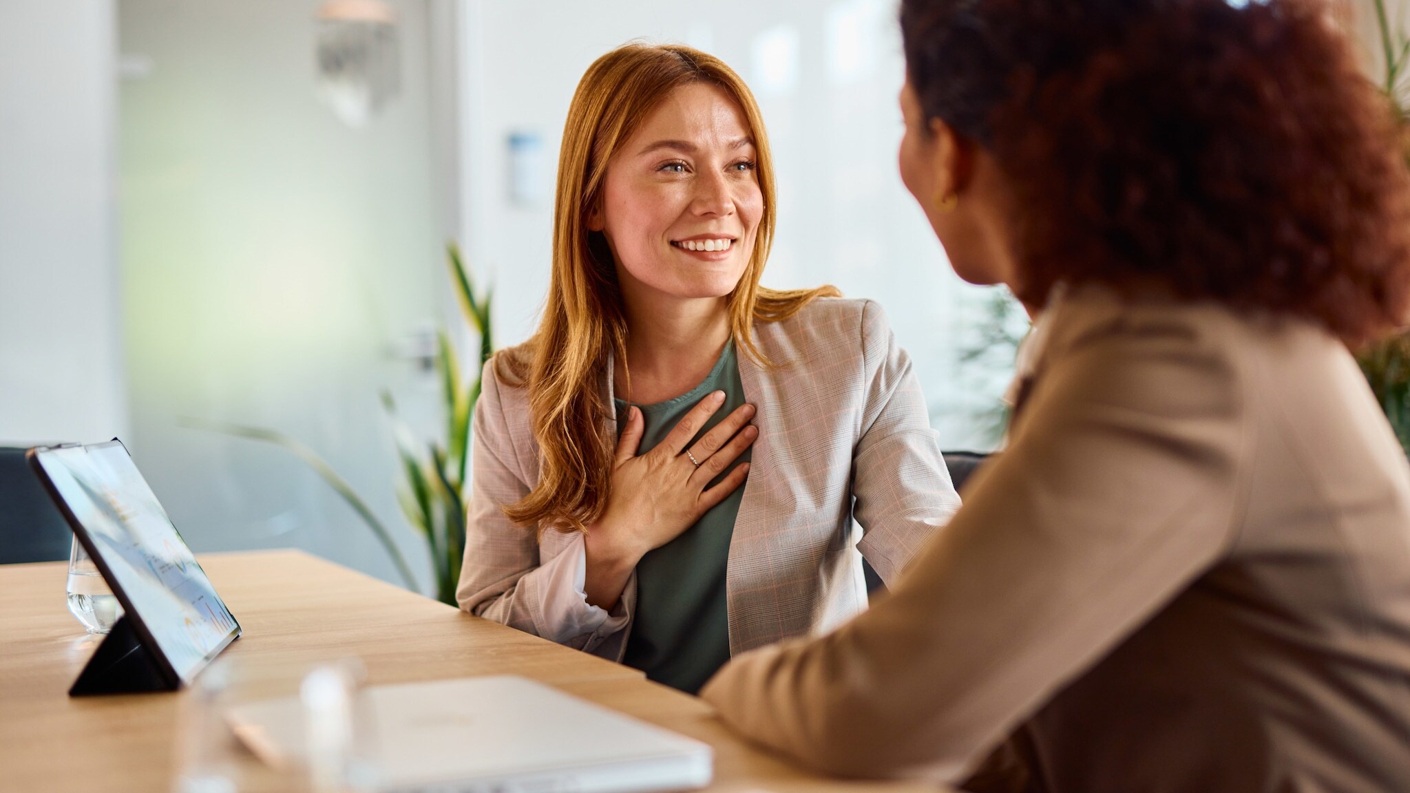 ine Frau im Blazer hat eine Hand auf die Brust gelegt und lächelt eine weitere Frau an, beide sitzen an einem Tisch, auf dem ein Tablet und ein Wasserglas stehen.