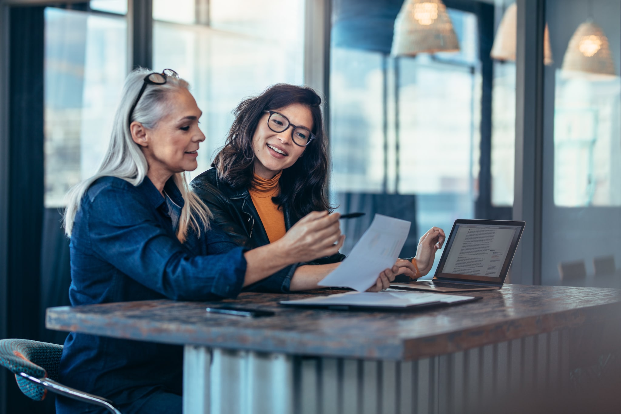 Zwei Frauen blicken auf Arbeitsunterlagen an einem Tisch mit Laptop in einem Büro.