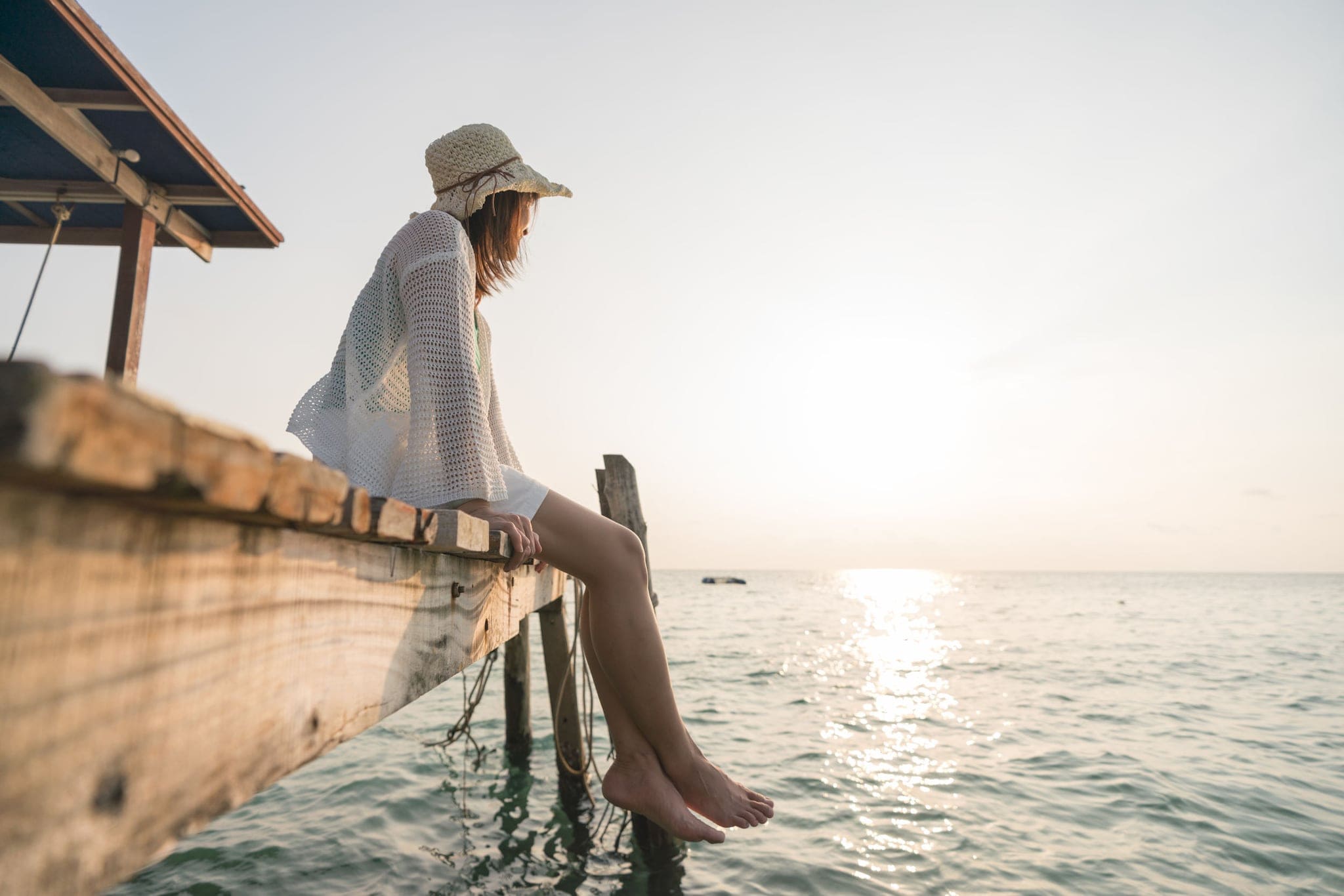 Frau mit Sonnenhut und barfuß, die auf einem Holzsteg sitzt und aufs Wasser schaut. Frau mit Sonnenhut und barfuß, die auf einem Holzsteg sitzt und aufs Wasser schaut.